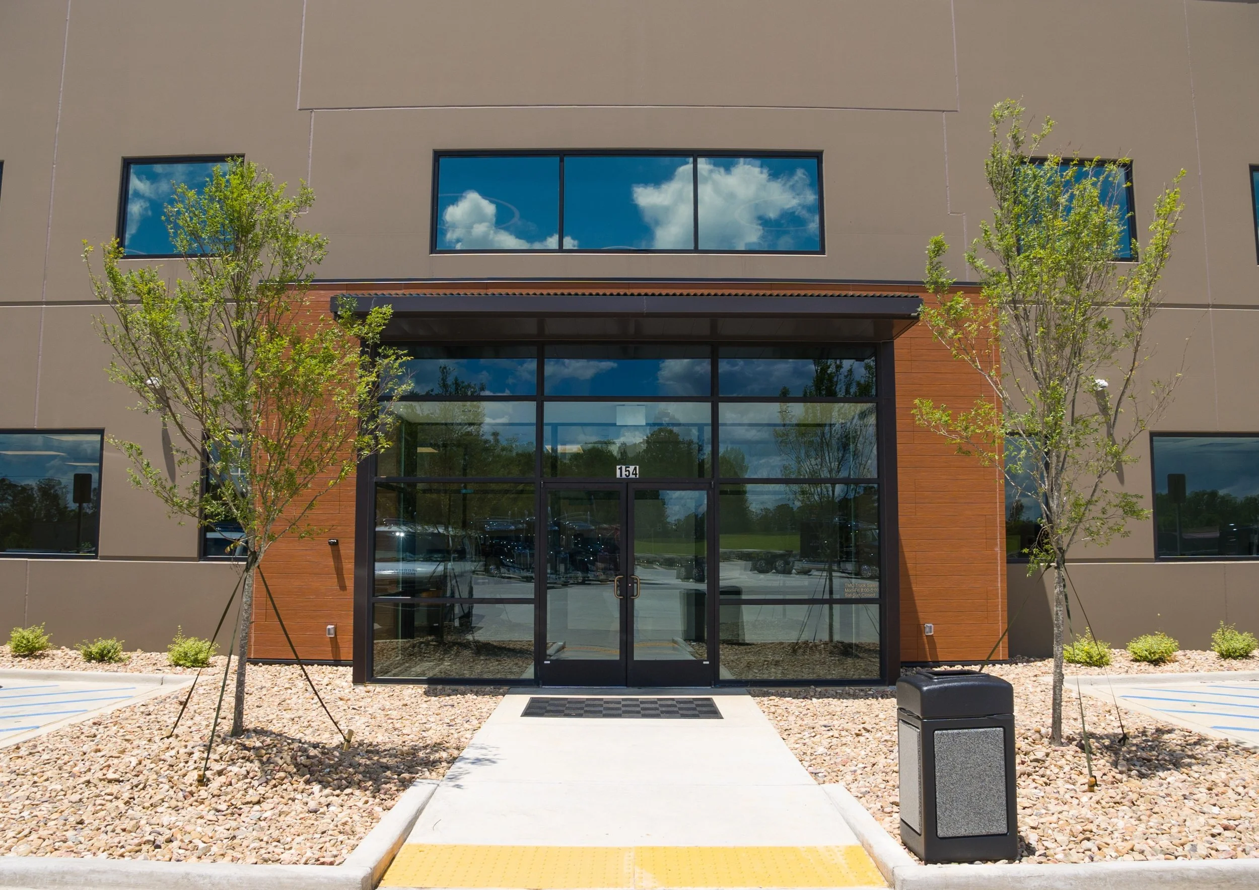 The image shows the front entrance of a modern building with large glass doors, a small concrete walkway, and two small trees flanking the entrance. The building has a beige exterior with wood accents surrounding the entrance. There are windows refle