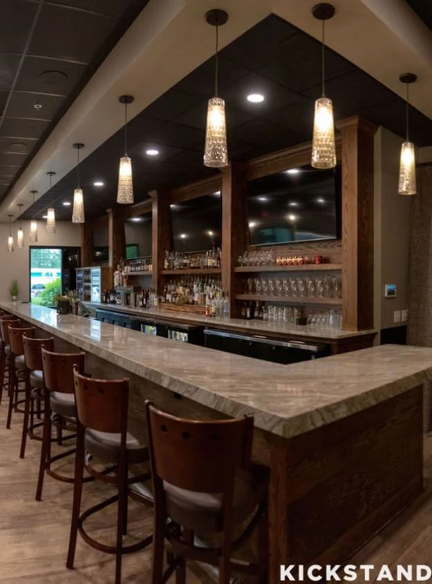 Interior of a modern bar with a long marble countertop, wooden bar stools, hanging pendant lights, and shelves stocked with various bottles and glassware.