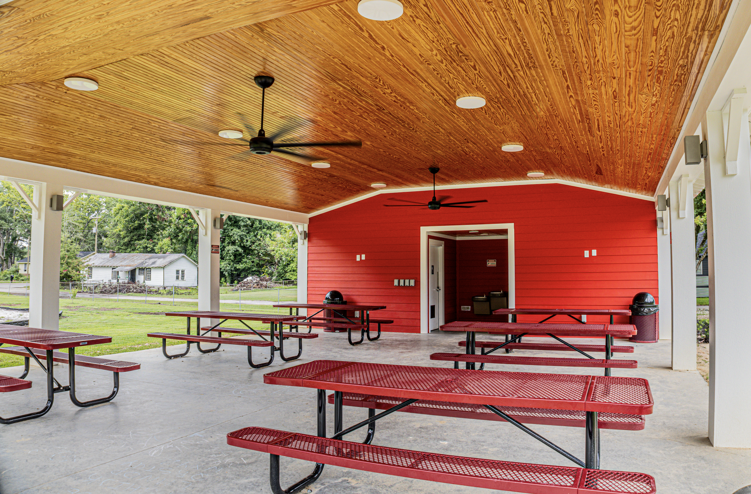 Covered outdoor picnic area with red benches, red tables, a red building, and a wooden ceiling with ceiling fans and lights.