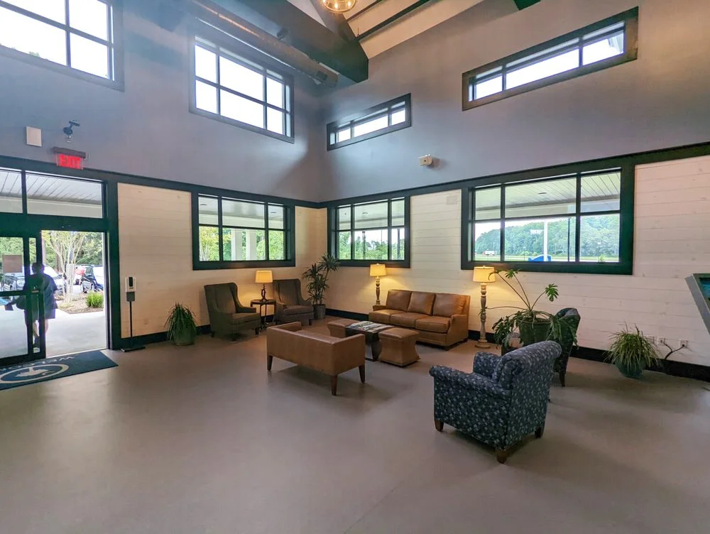 A spacious waiting area with a beige leather sofa, two patterned armchairs, several side tables with lamps, and large windows letting in natural light. The room has high ceilings with exposed beams and a view outside with greenery.