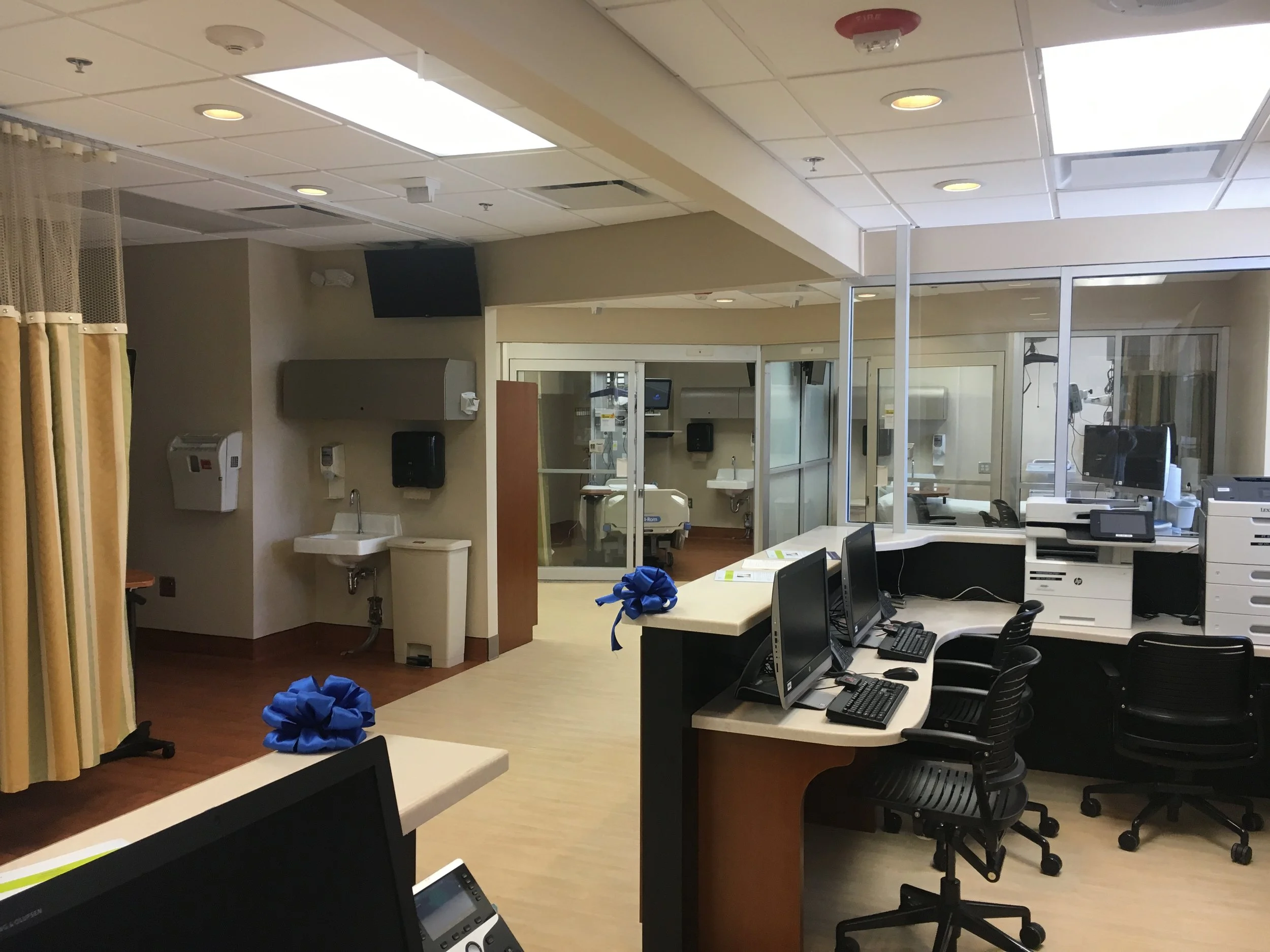 A hospital room with multiple computer workstations, medical equipment, and a glass partition, decorated with blue ribbons and bows.