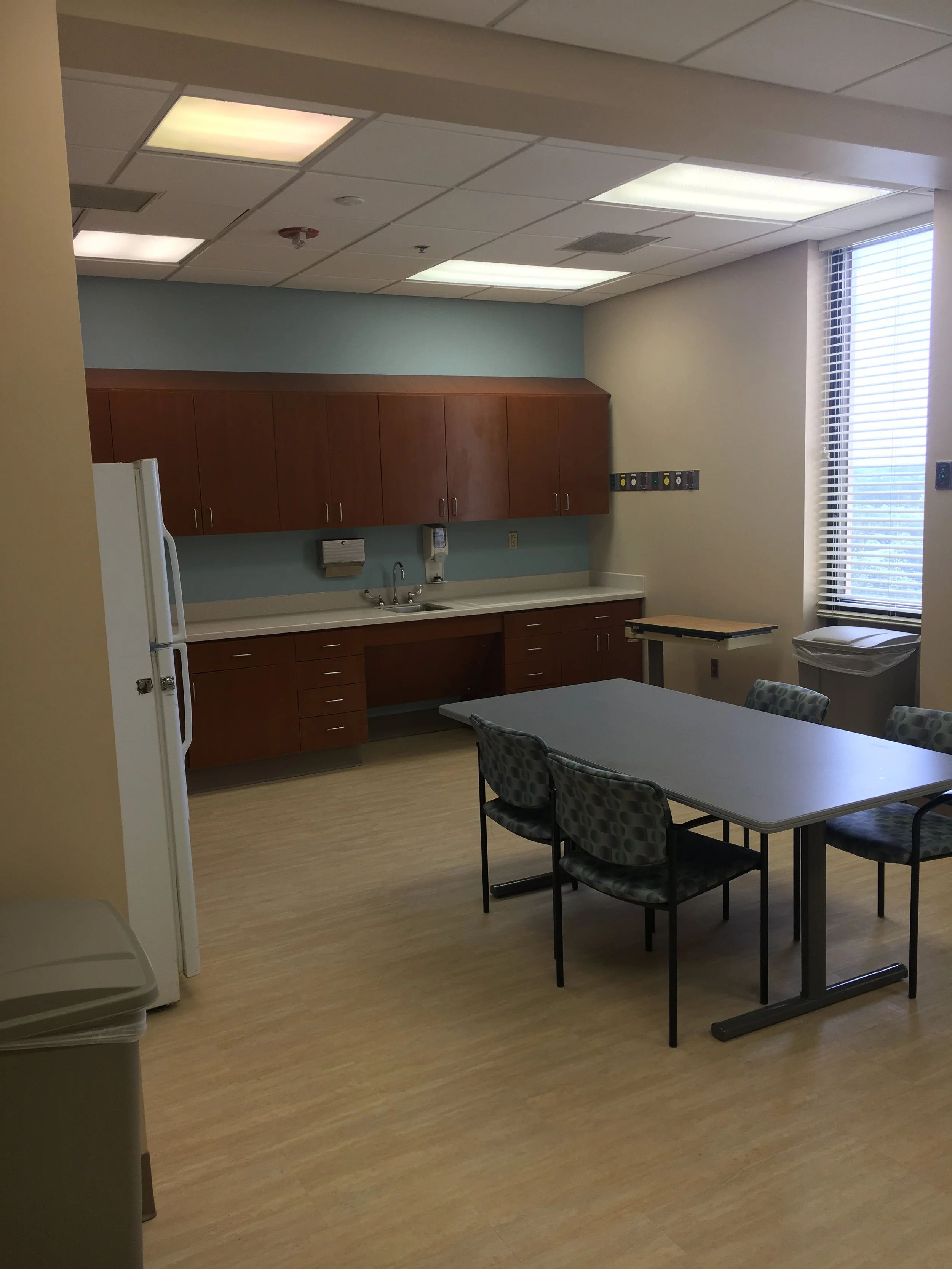 A kitchenette in a room with wooden cabinets, a sink, a wall-mounted telephone, and a window with blinds. There are tables and chairs in the room.