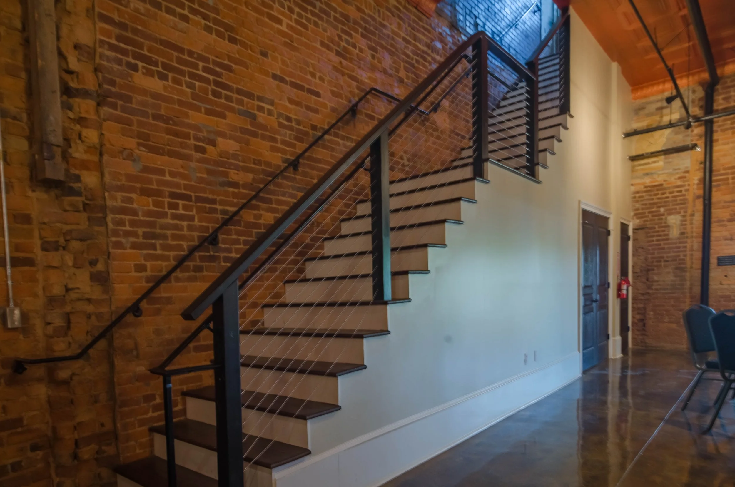 Interior view of an industrial-style space with exposed brick walls, a modern staircase with black metal frame, wooden steps, and cable railing, and a high wooden ceiling with visible pipes and beams.