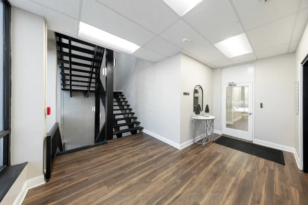 Empty modern lobby area with dark wood flooring, white walls, a small table with decorative items, a glass door, and a black staircase leading upstairs.