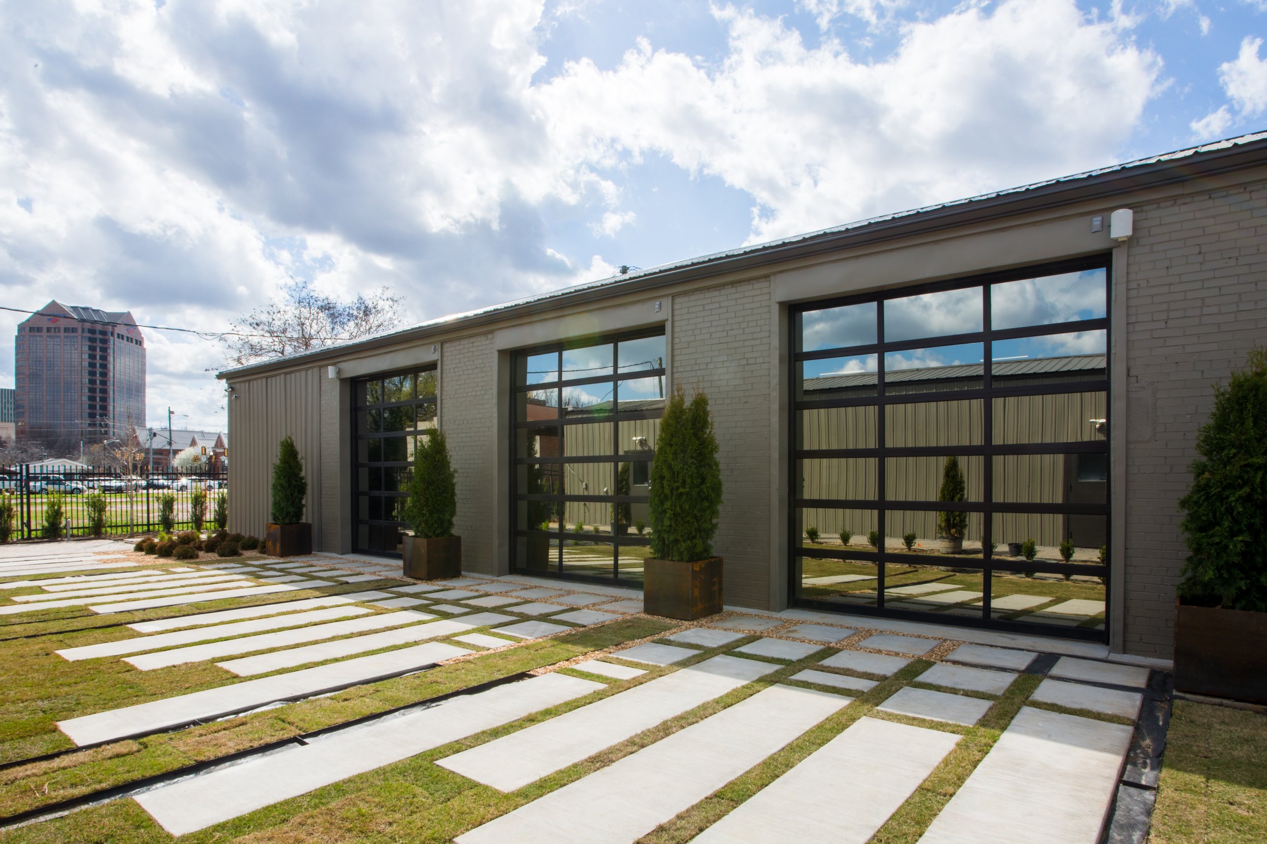 Exterior of a modern building with large glass windows, potted tall green plants, and a pathway with white rectangular stones set in grass, under a partly cloudy sky.