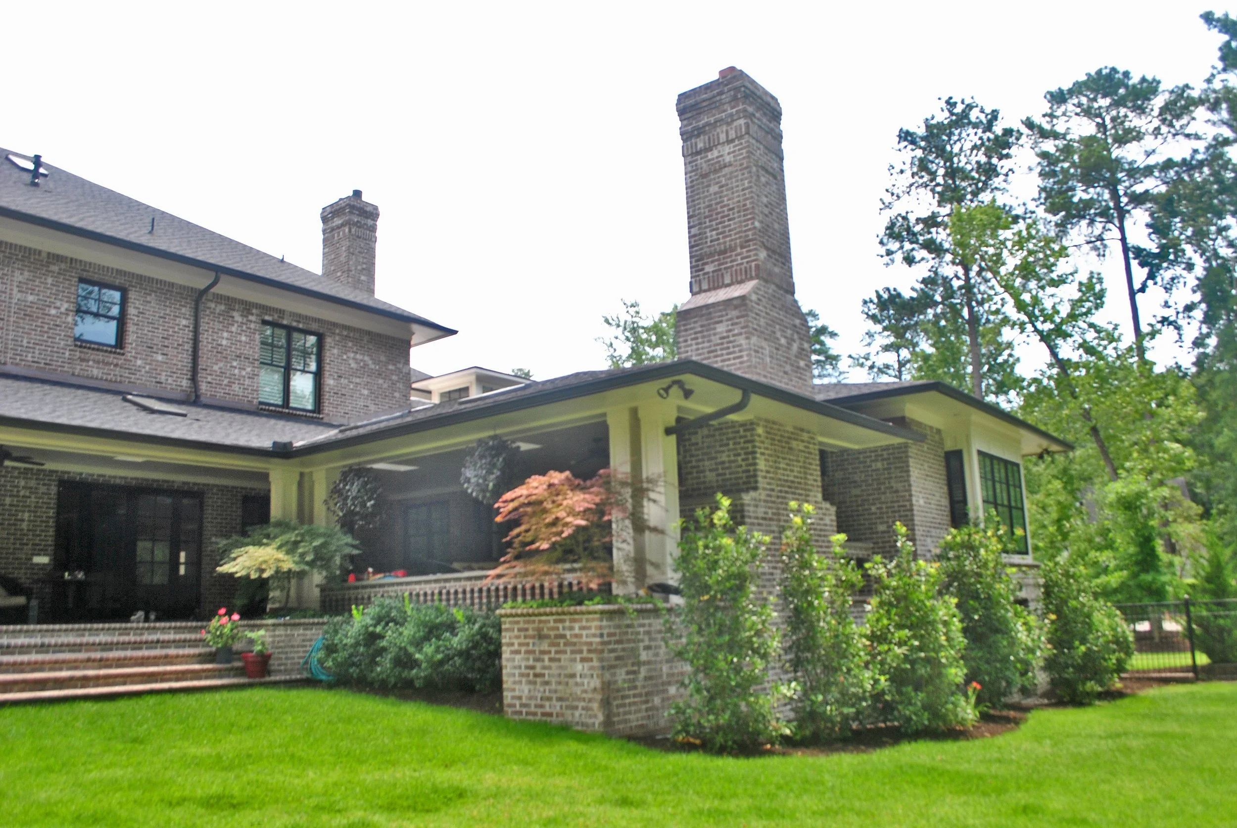 A two-story brick house with a backyard, green lawn, steps, and landscaped garden, surrounded by tall trees.