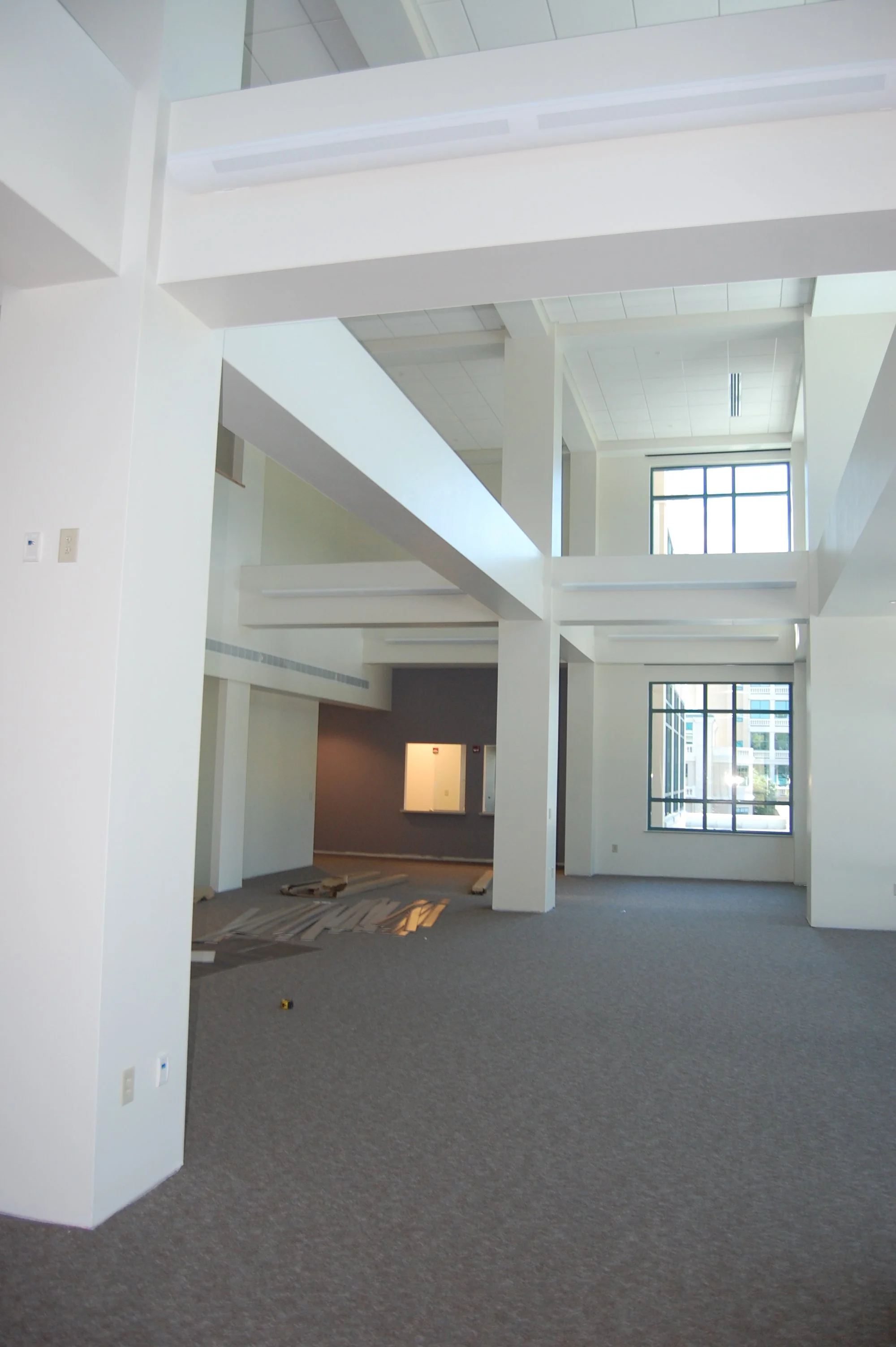 Interior of a modern, spacious building with high ceilings, large windows, and gray carpeted floors. There are white walls and columns, with some construction materials on the floor, indicating ongoing work or recent renovations.