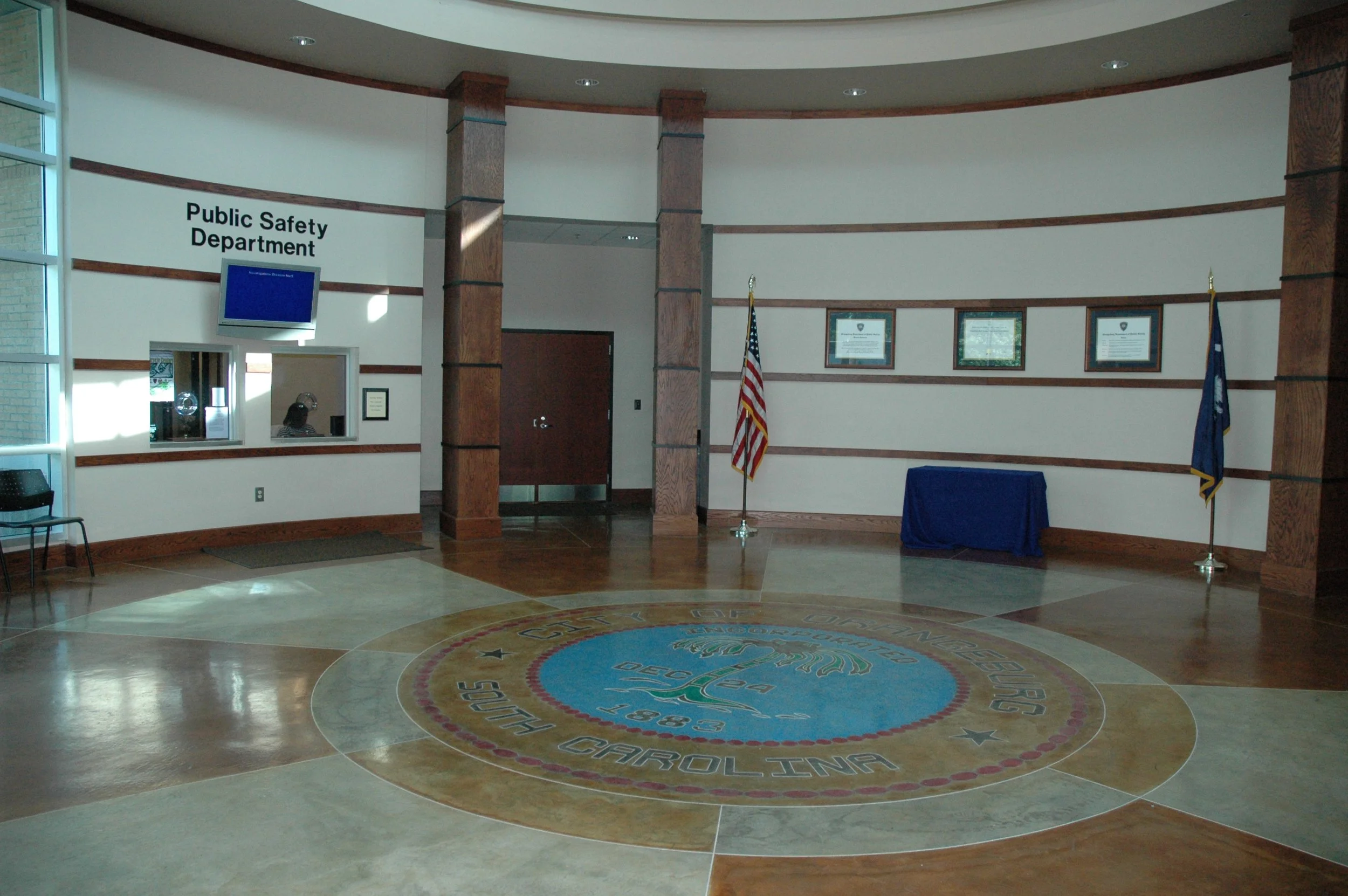 Interior of a government building with a circular floor design featuring the seal of North Carolina and words 'Government of North Carolina'. There are framed documents on the wall, an American flag and a state flag, and a blue table with cloth.