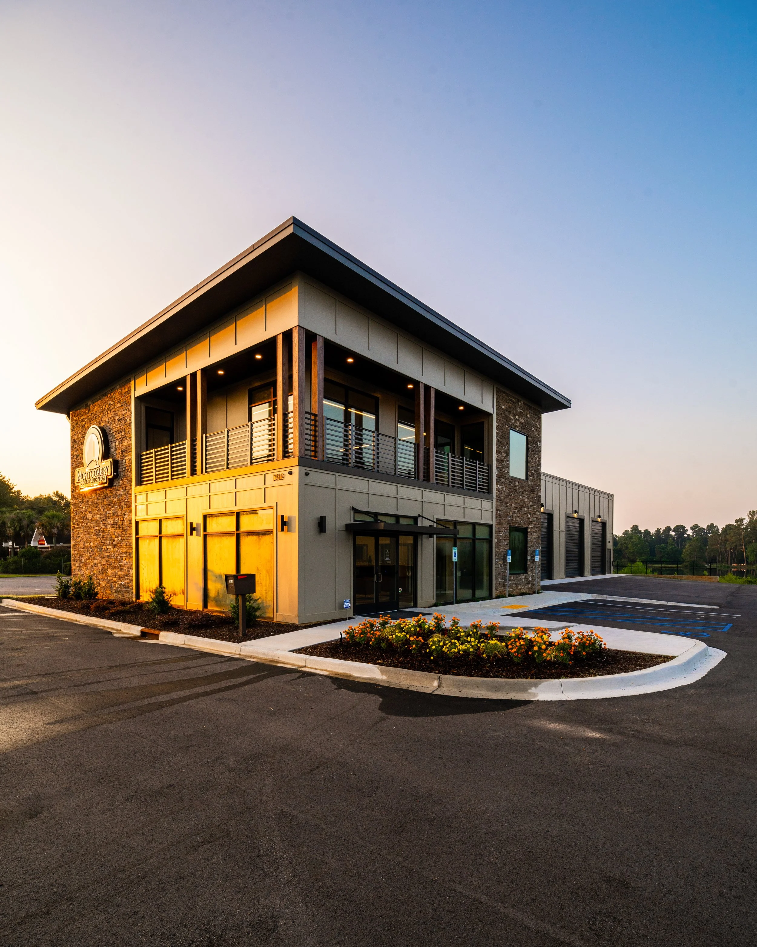 Modern commercial building with brick and siding exterior, large windows, and a balcony, located in a parking lot during sunset, with landscaped flower beds and parking spaces.