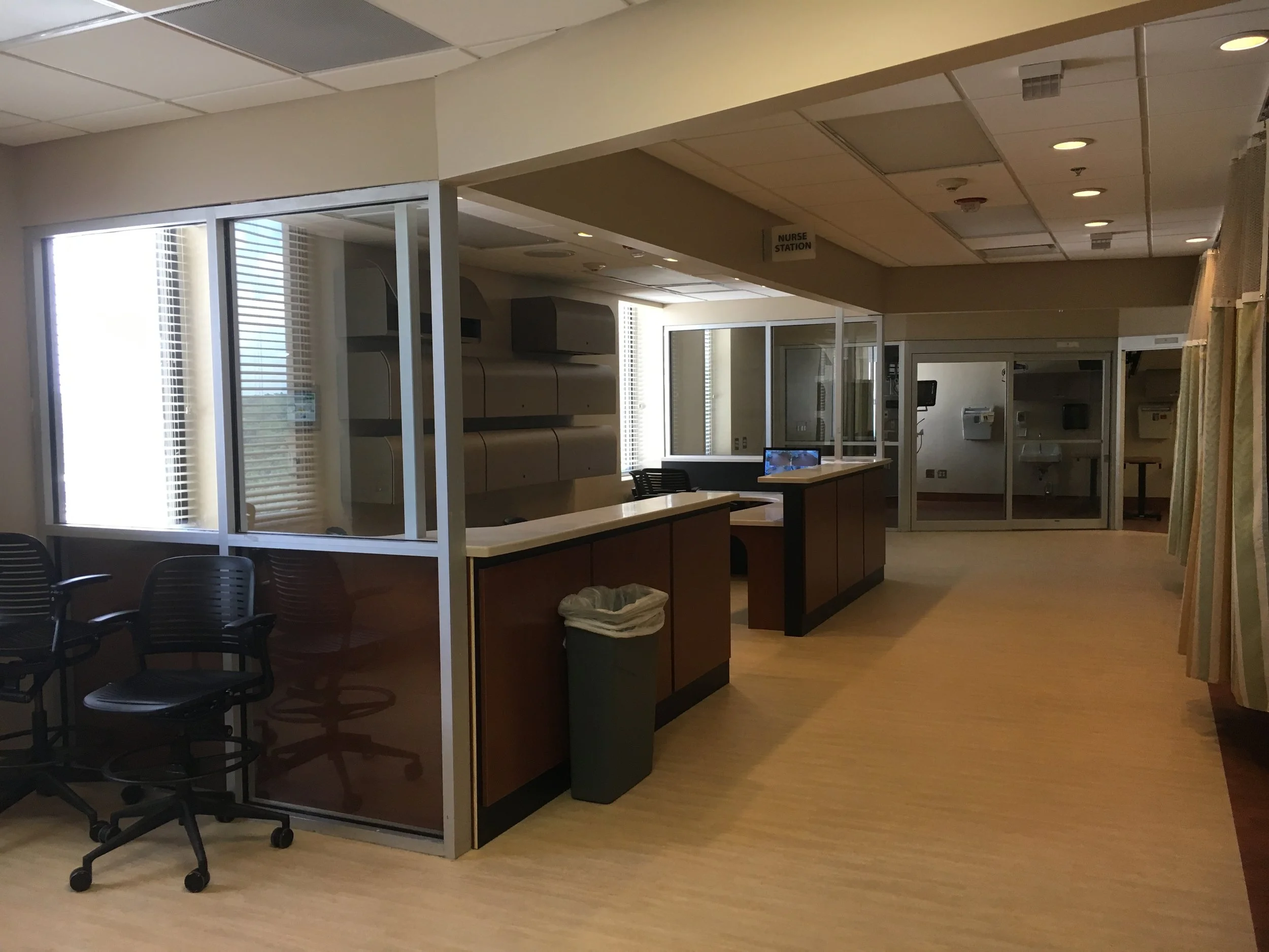 View of a hospital or clinic waiting area with chairs, a nurse station, and medical equipment in a well-lit room.