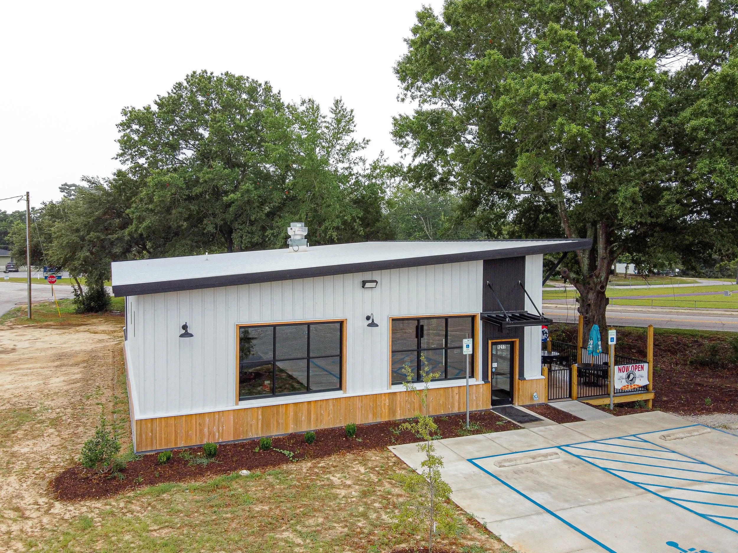 Exterior of a small modern restaurant or shop with large glass windows and a white and wood panel facade, situated in an area with trees and a parking lot.