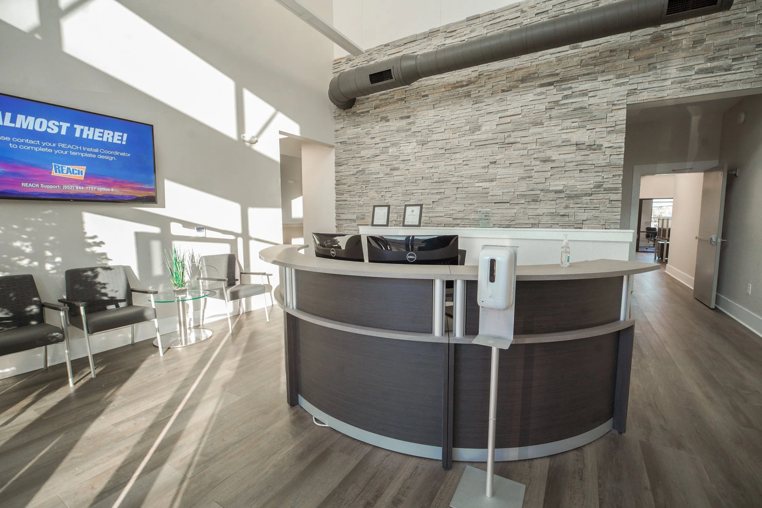 Reception area with a curved front desk, black computer, hand sanitizer, framed certificates, chairs, a large wall mounted television screen displaying a message about reaching out to a support contact, stone accent wall, and visible ventilation duct