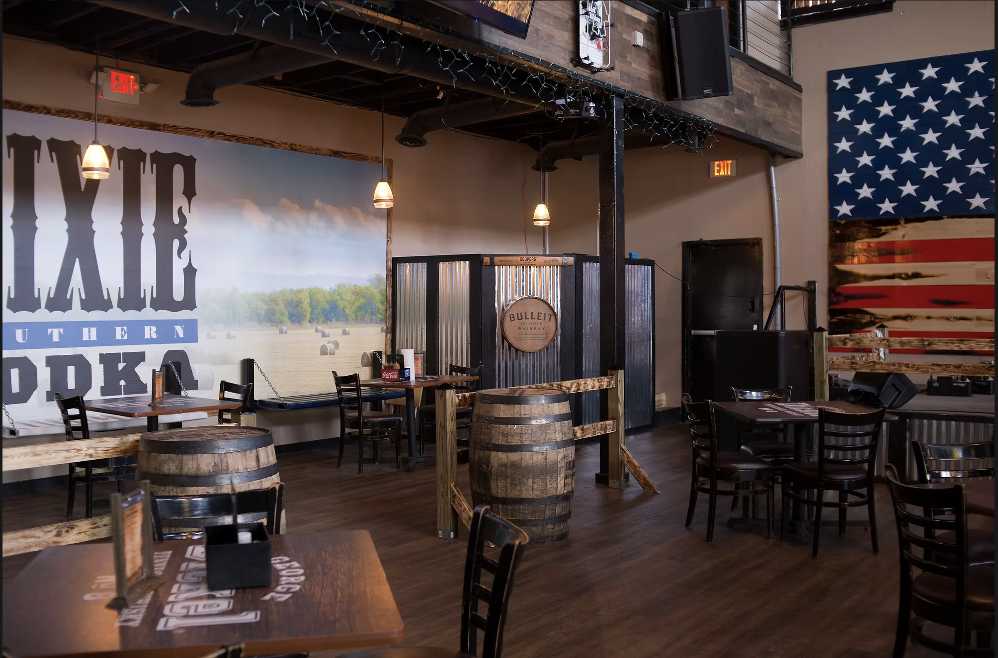 Interior of a rustic-style bar or restaurant with wooden tables, chairs, and barrel decorations. There is a large American flag-themed wooden panel on the wall. The space has hanging pendant lights, a hardware metallic bar counter, and rustic decor e