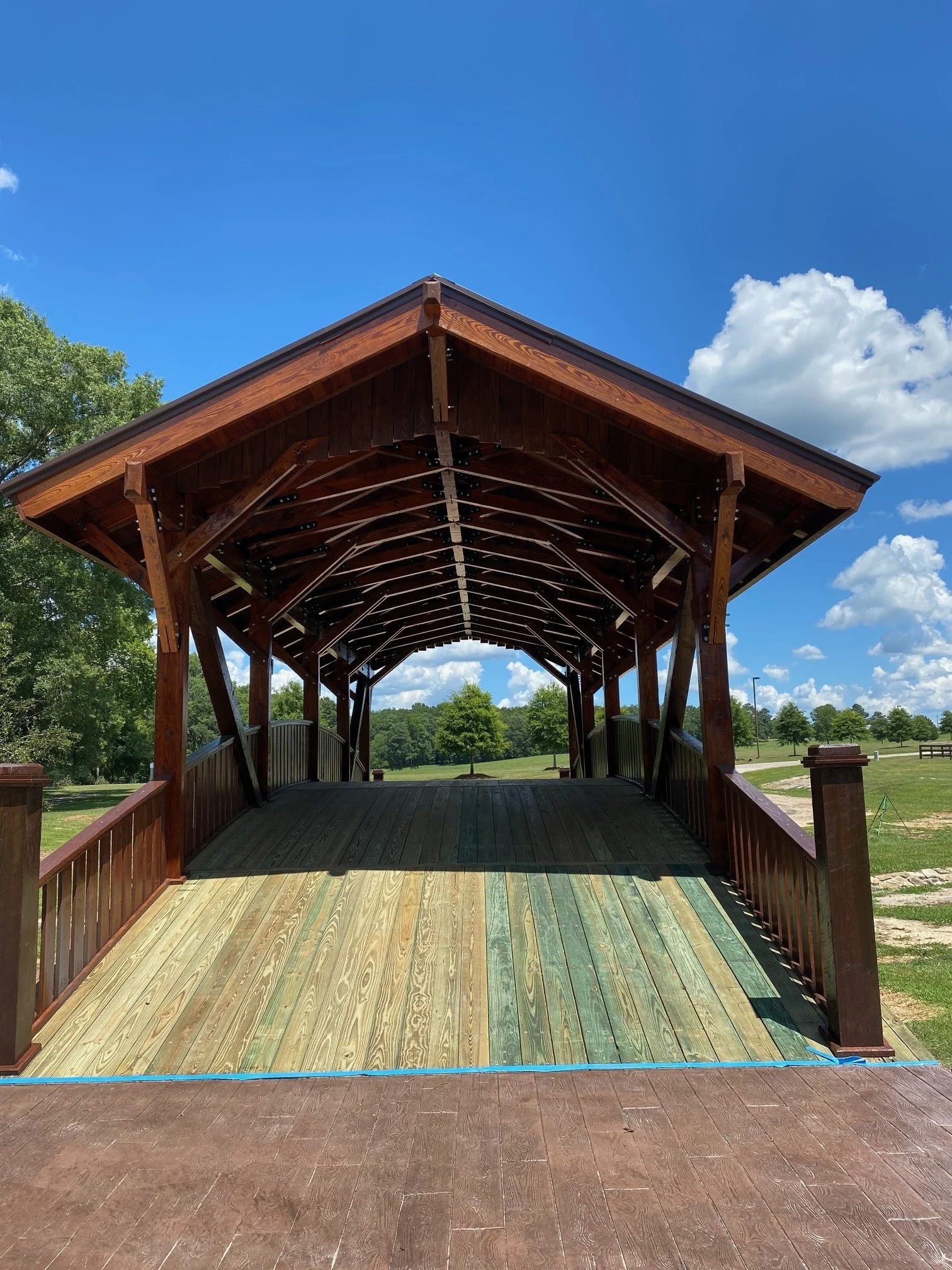 A wooden pedestrian bridge with a roof under a bright blue sky with some clouds, surrounded by green trees and open grassy areas.