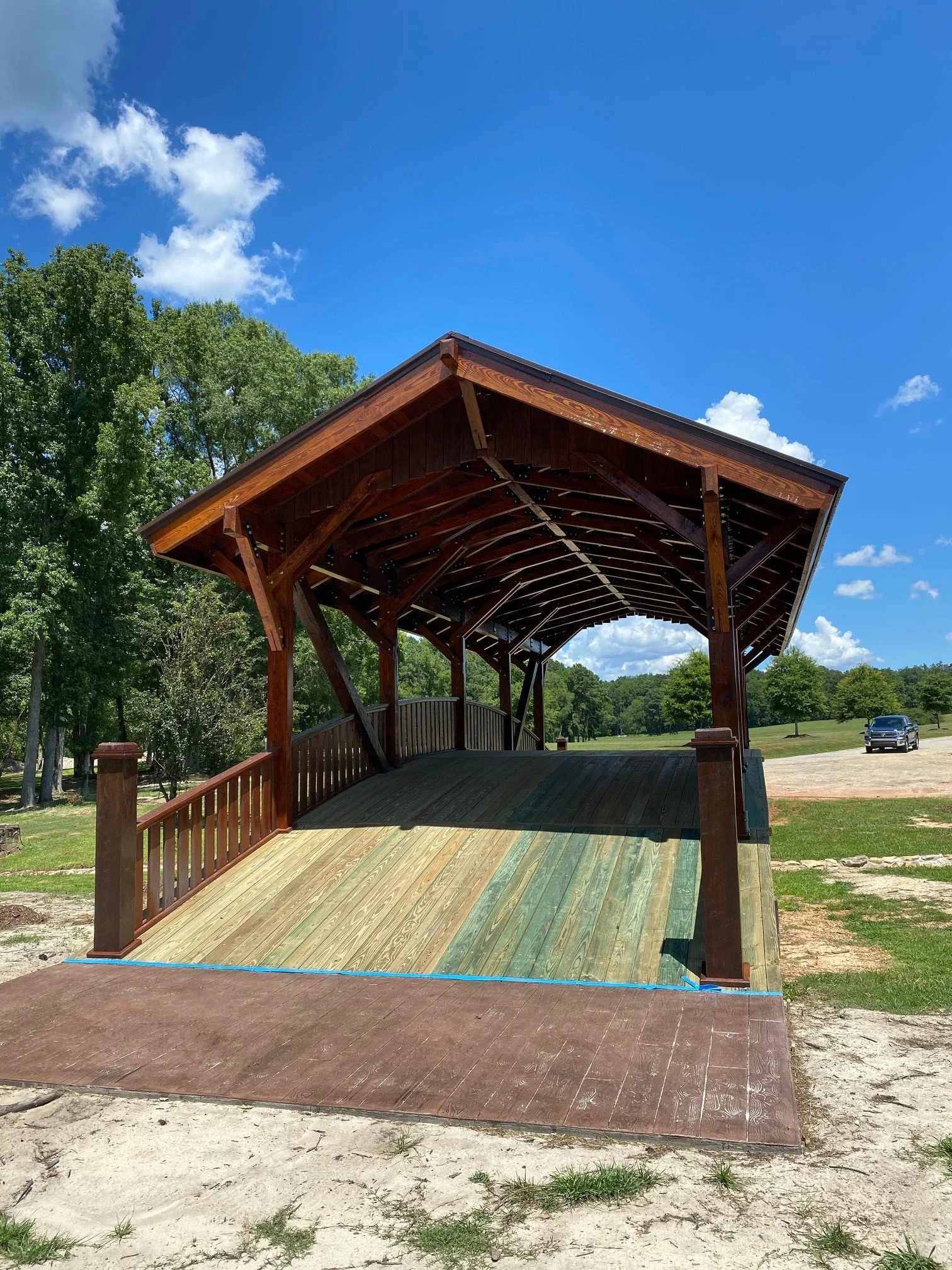 A wooden pavilion with a sloped roof and open sides set in a park with green trees and a grassy field in the background. A black vehicle is parked on a dirt and gravel area nearby.
