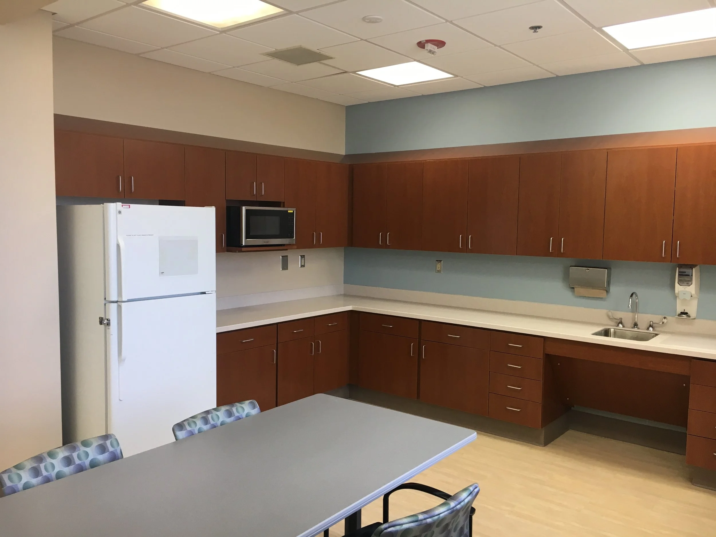 An office break room with wooden cabinets, a white refrigerator, a microwave oven, a sink, and a table with chairs.