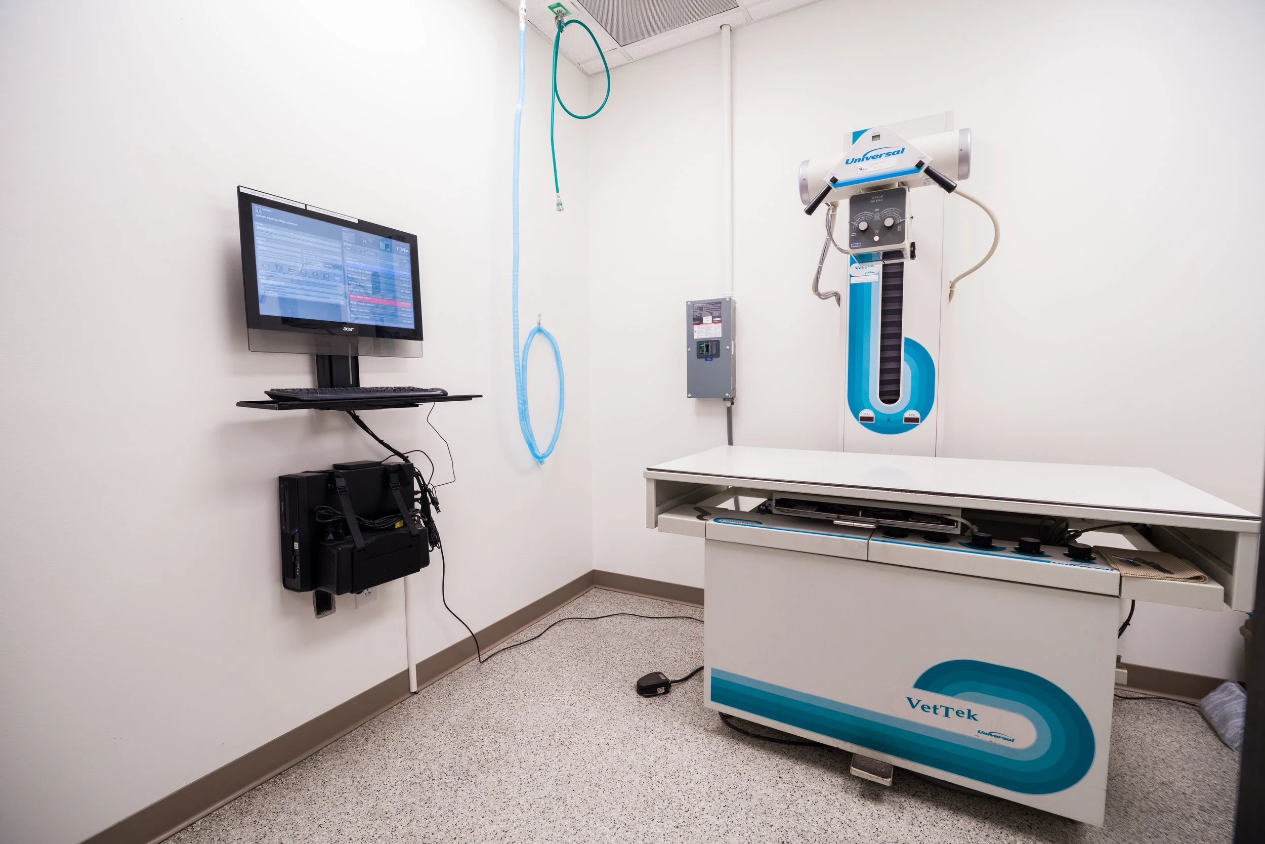 An examination room with a veterinary X-ray machine, a computer monitor, and various medical equipment against white walls and a tiled floor.