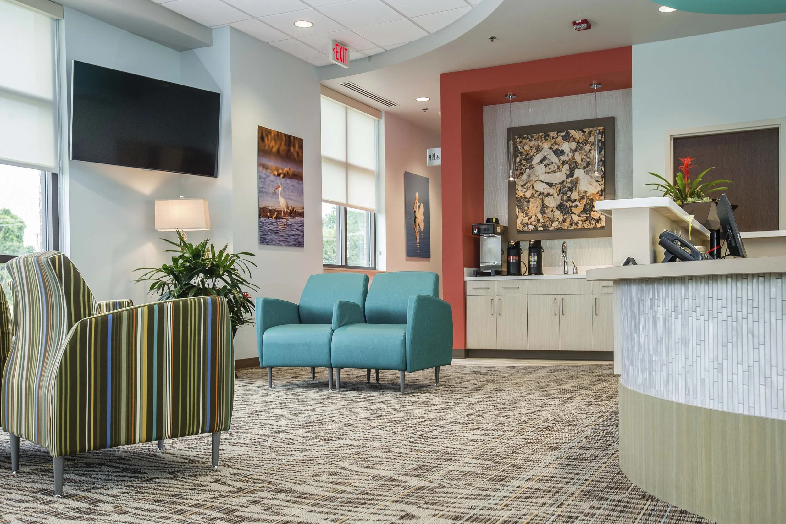 Lobby area with striped armchairs, teal sofa, potted plants, wall art, a TV, and a reception desk with a plant and computer.