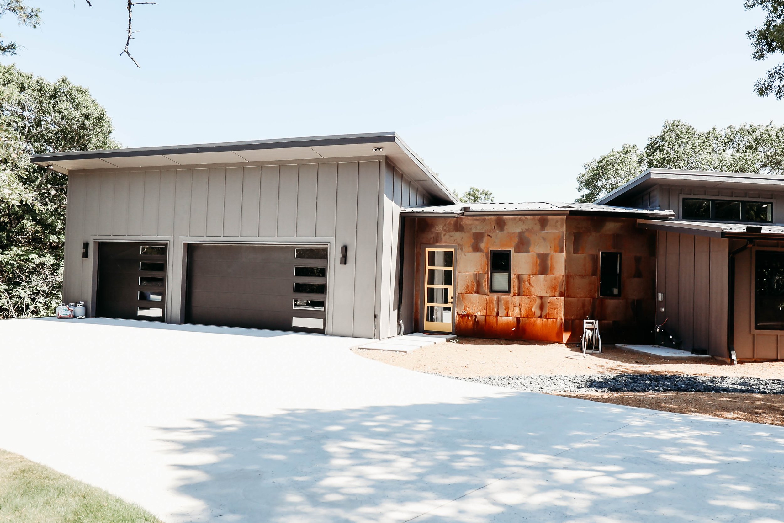 Modern house with gray and rust-colored exterior walls, large garage doors, and surrounding trees under a clear sky.
