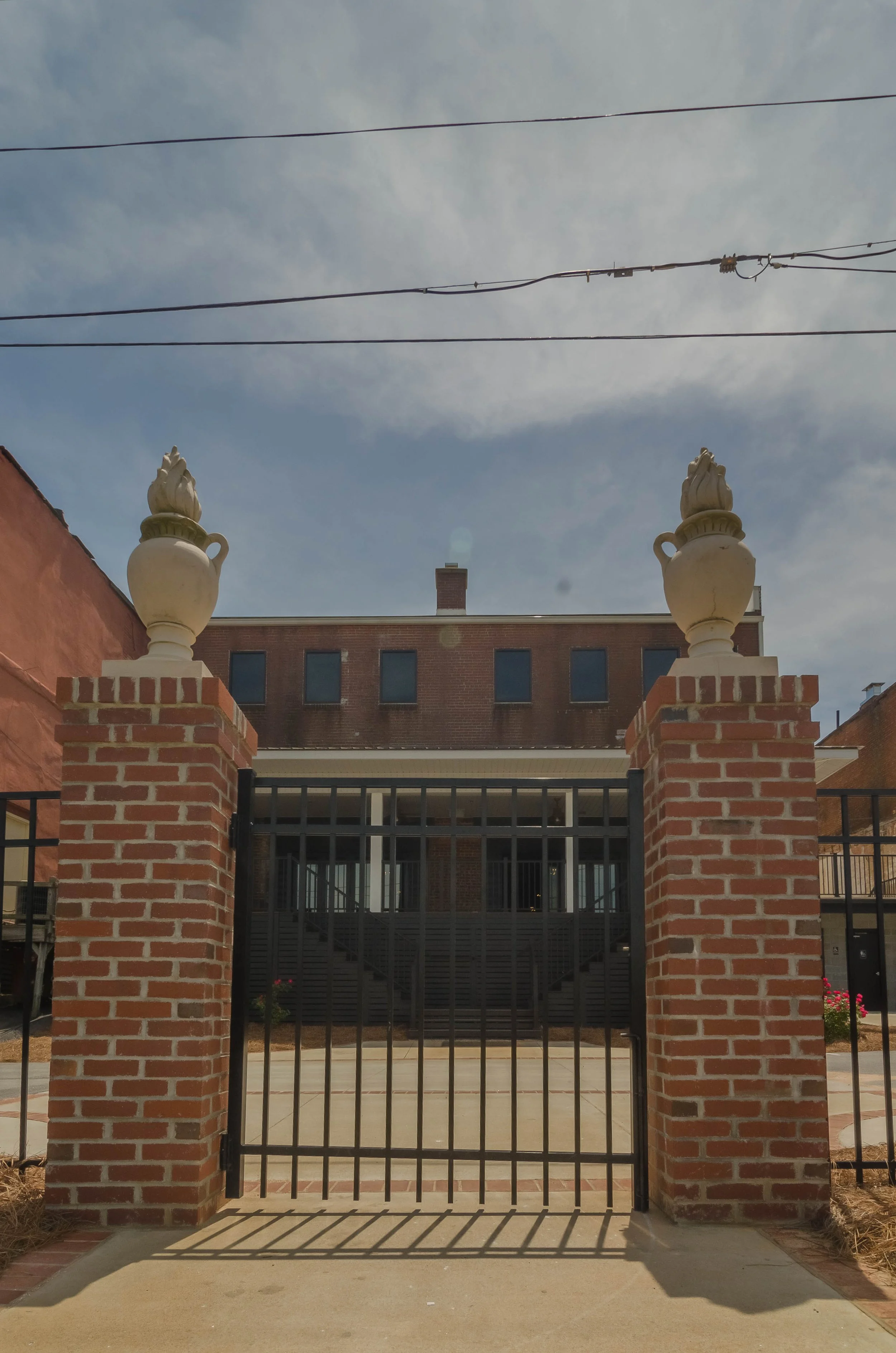 Brick pillars with decorative urns on top, black metal gate, brick building in background, sky with clouds, electrical wires overhead.
