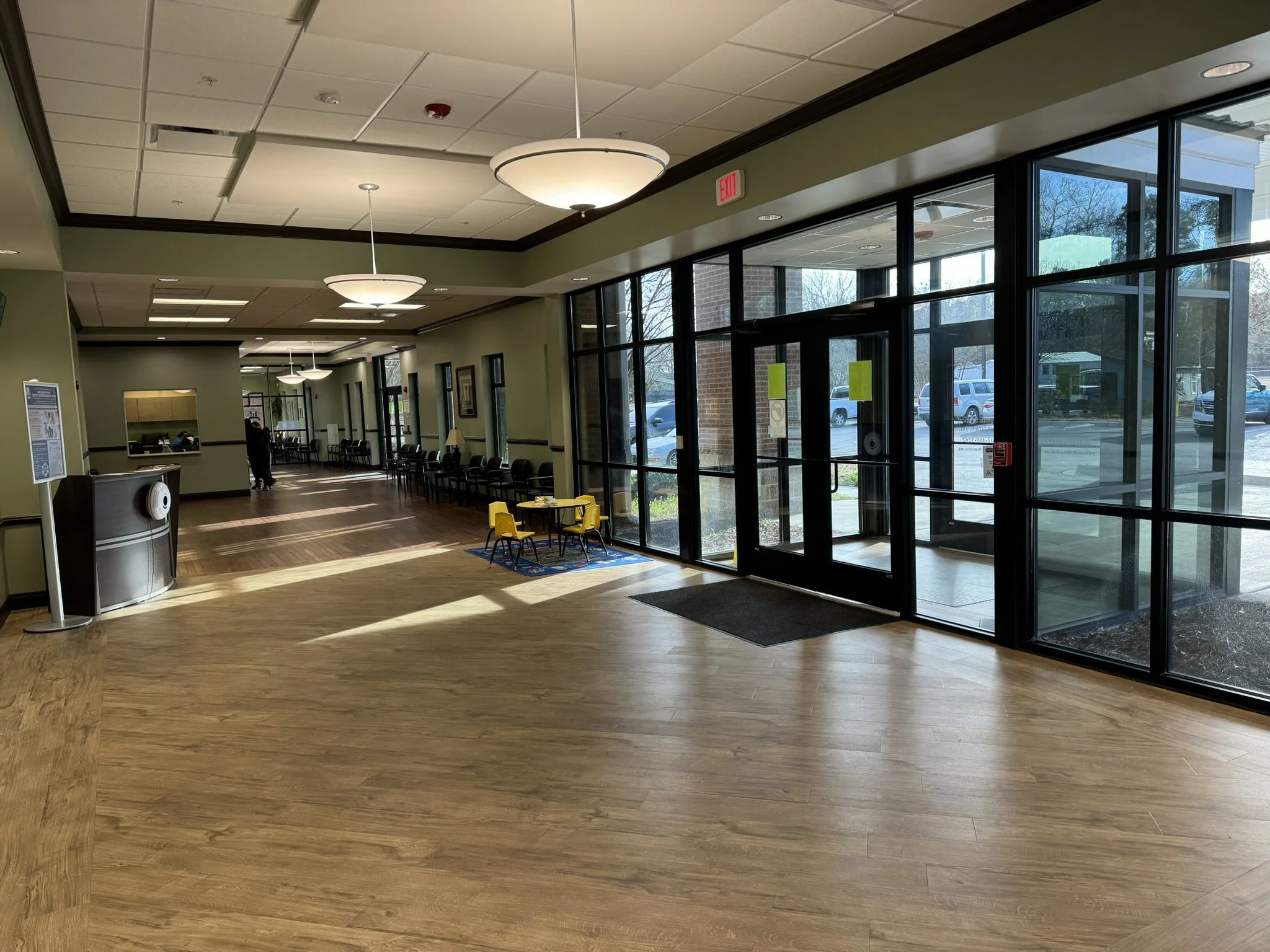 Interior view of a hospital waiting area with a row of black chairs, large glass entrance doors, and ceiling lights. Outside, there are cars visible.