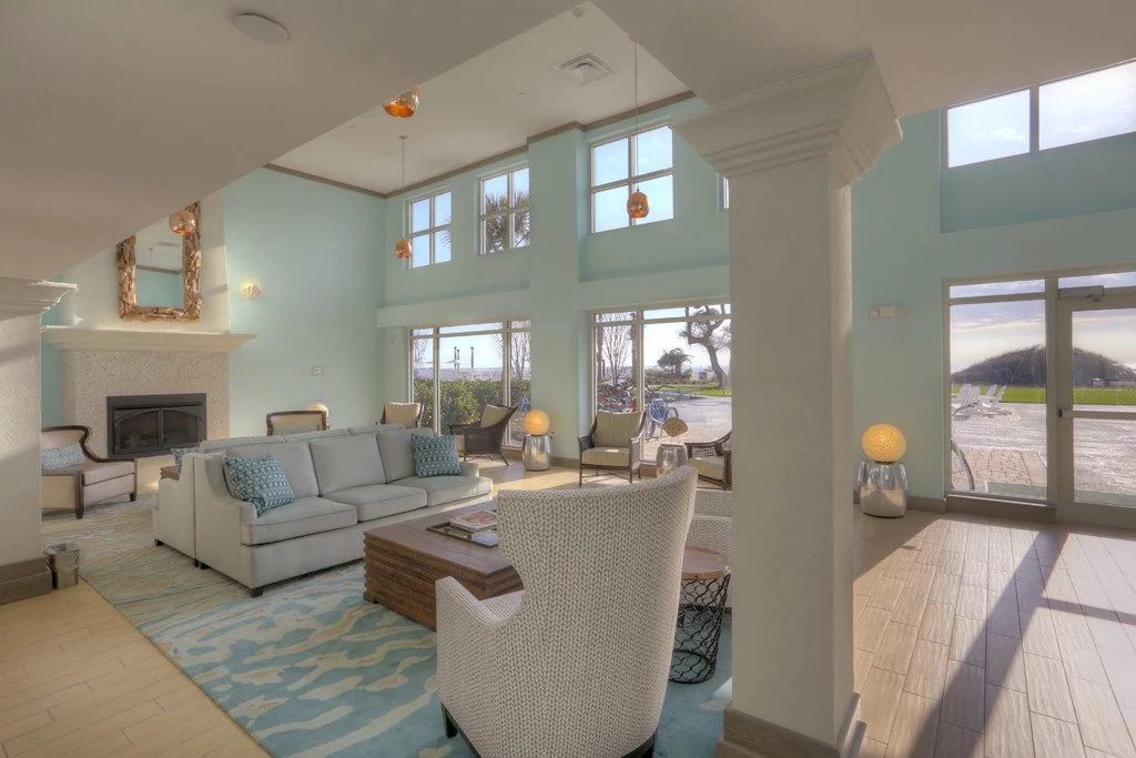 Indoor lounge area with light blue walls, large windows, and white furniture including a sofa and armchairs. There is a fireplace, ceiling lights, and natural light streaming in. Outside, a parking lot and landscape are visible through the windows.