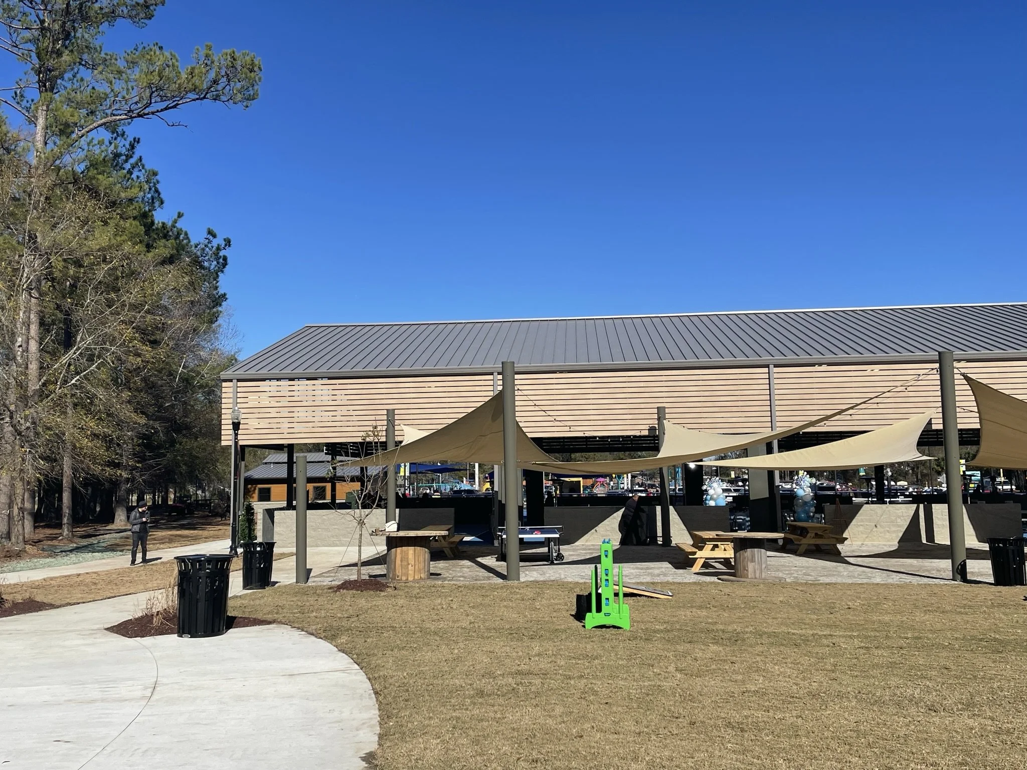 An outdoor public space with a large open grassy area, a paved walkway, trees, and a modern building with a metal roof. There are shade sails over seating areas, trash cans, and a person walking. The sky is clear and blue.