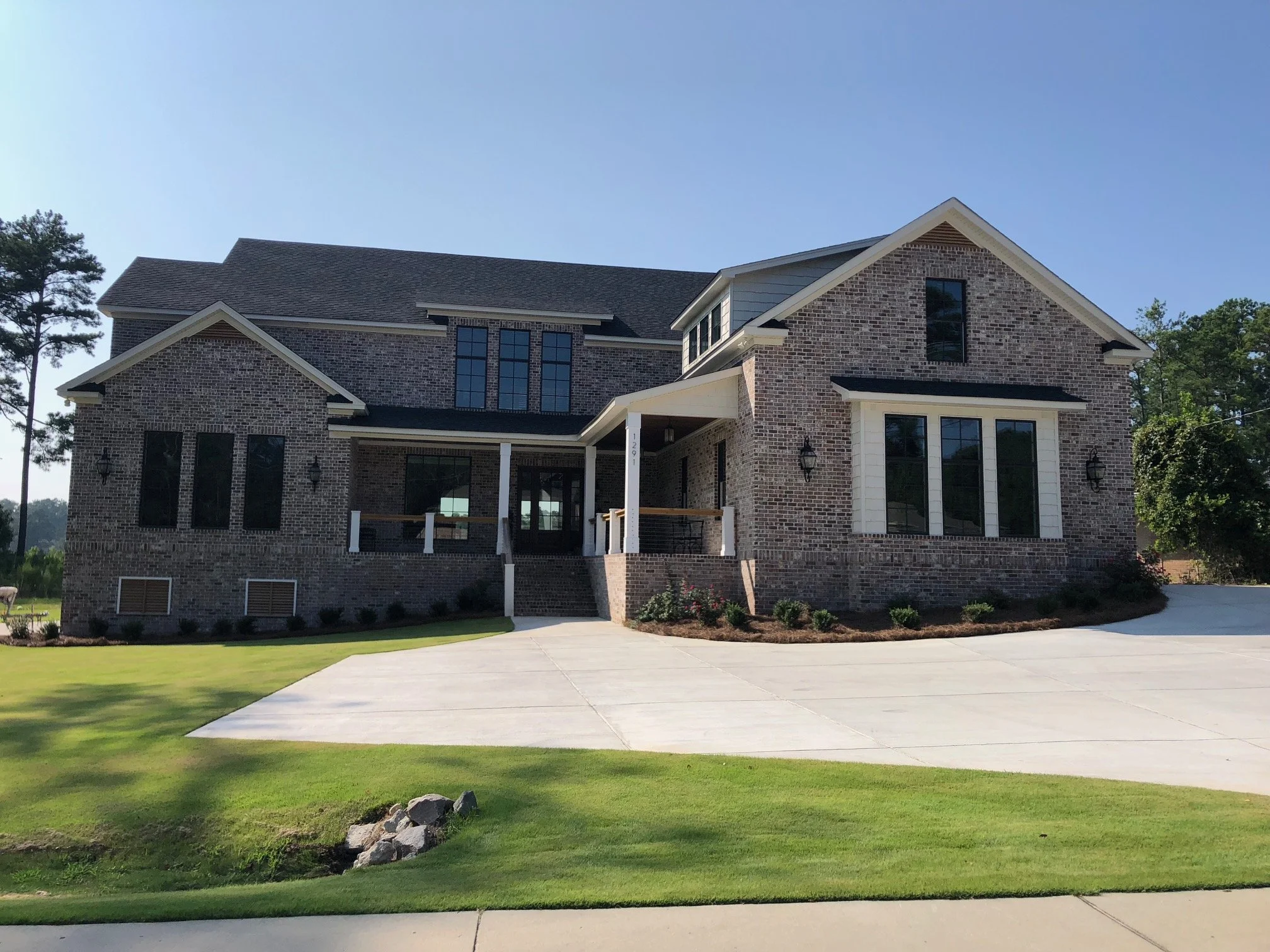 A large, modern brick house with multiple levels, large windows, and a front porch, surrounded by a well-maintained lawn and trees.