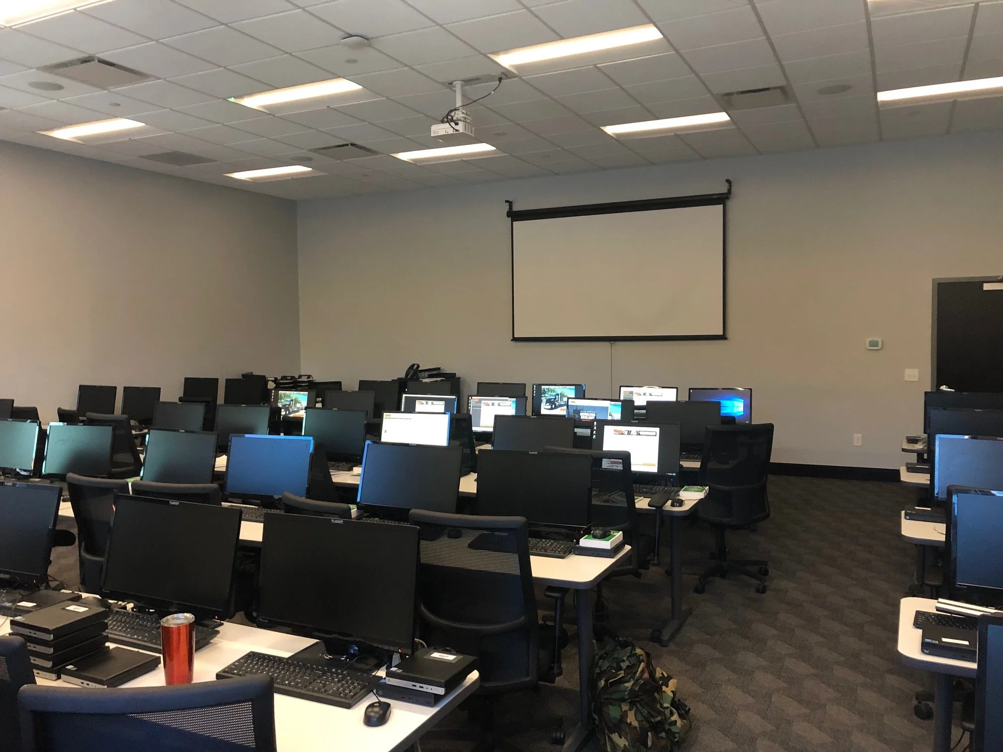 A classroom equipped with numerous computers on white tables, arranged in rows, with black chairs. The room has a drop ceiling with fluorescent lights, and a projection screen hangs on the front wall. There is a control panel on the right side of the