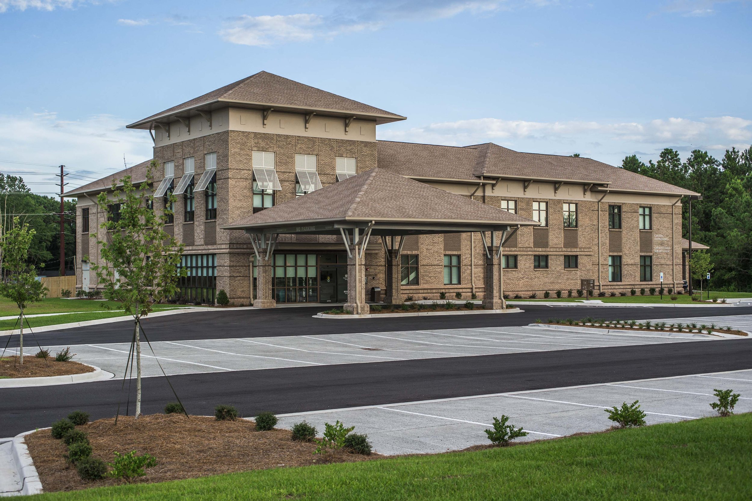 A modern multi-story brick building with a covered entrance and several windows, surrounded by a freshly paved parking lot and landscaped greenery, under a partly cloudy sky.