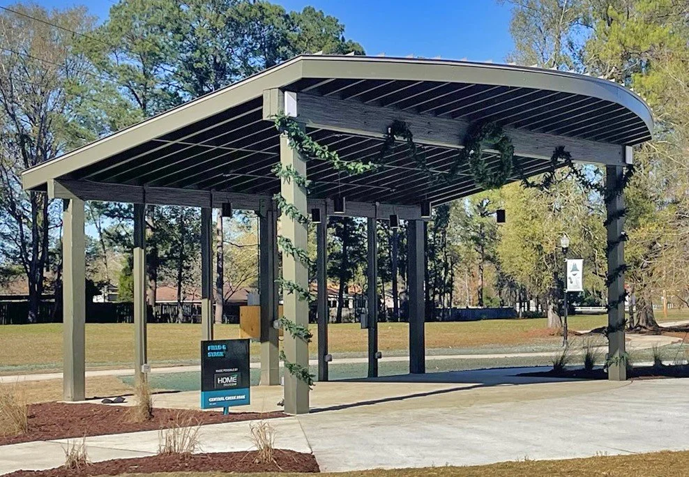 A covered outdoor public pavilion with string lights and greenery decorations, located in a park with trees and grass in the background.