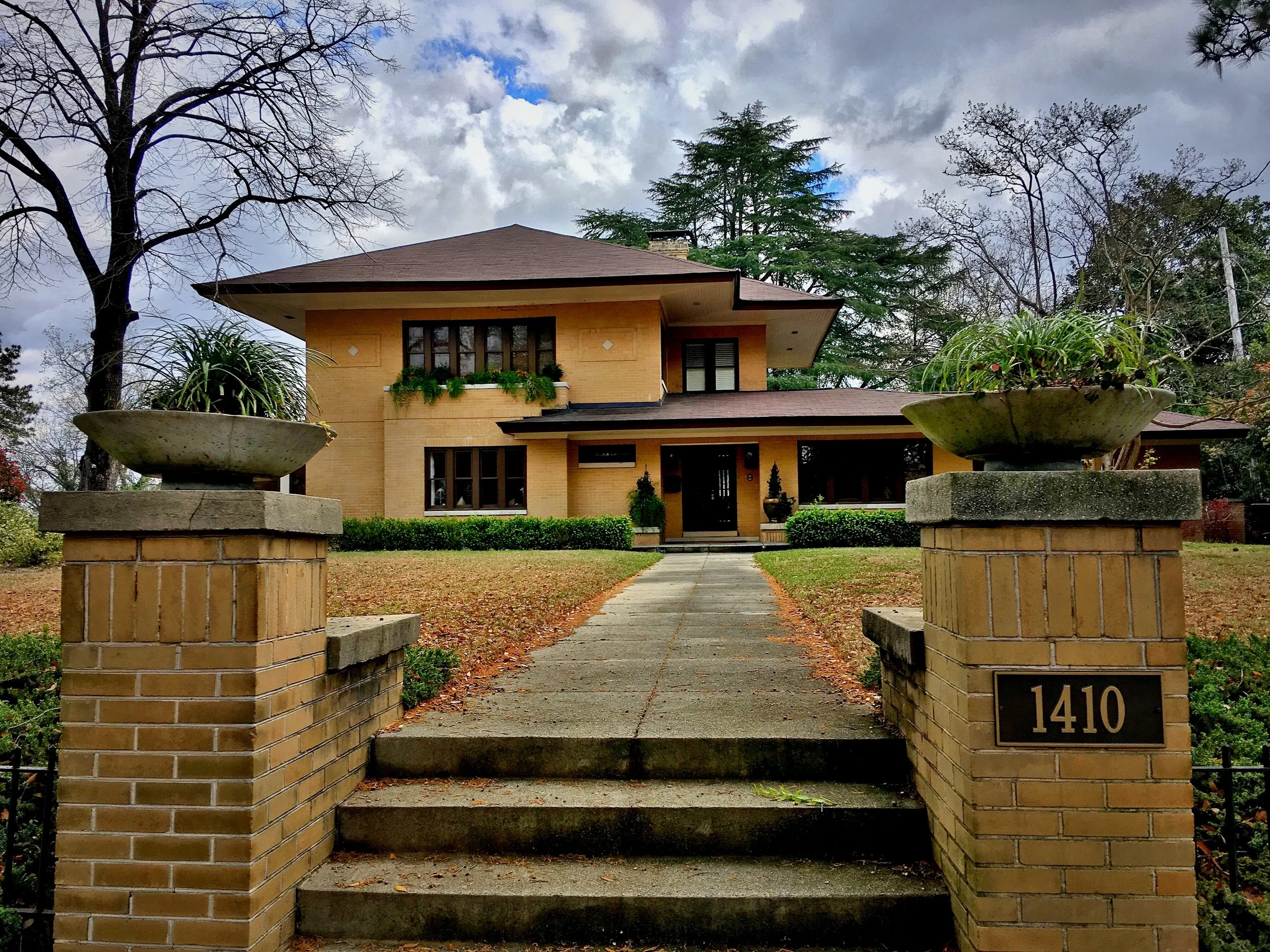 Front view of a yellow brick house surrounded by trees and shrubs, with a concrete walkway leading to the door and a decorative brick fence with two large flower pots on either side.