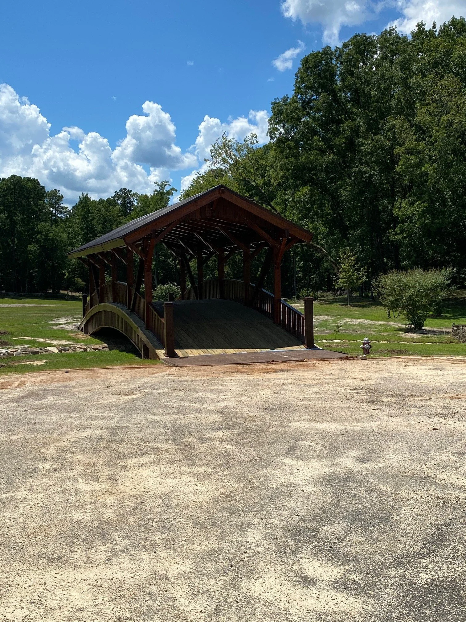 A wooden arched bridge in a park with green grass, trees, and a partly cloudy sky.