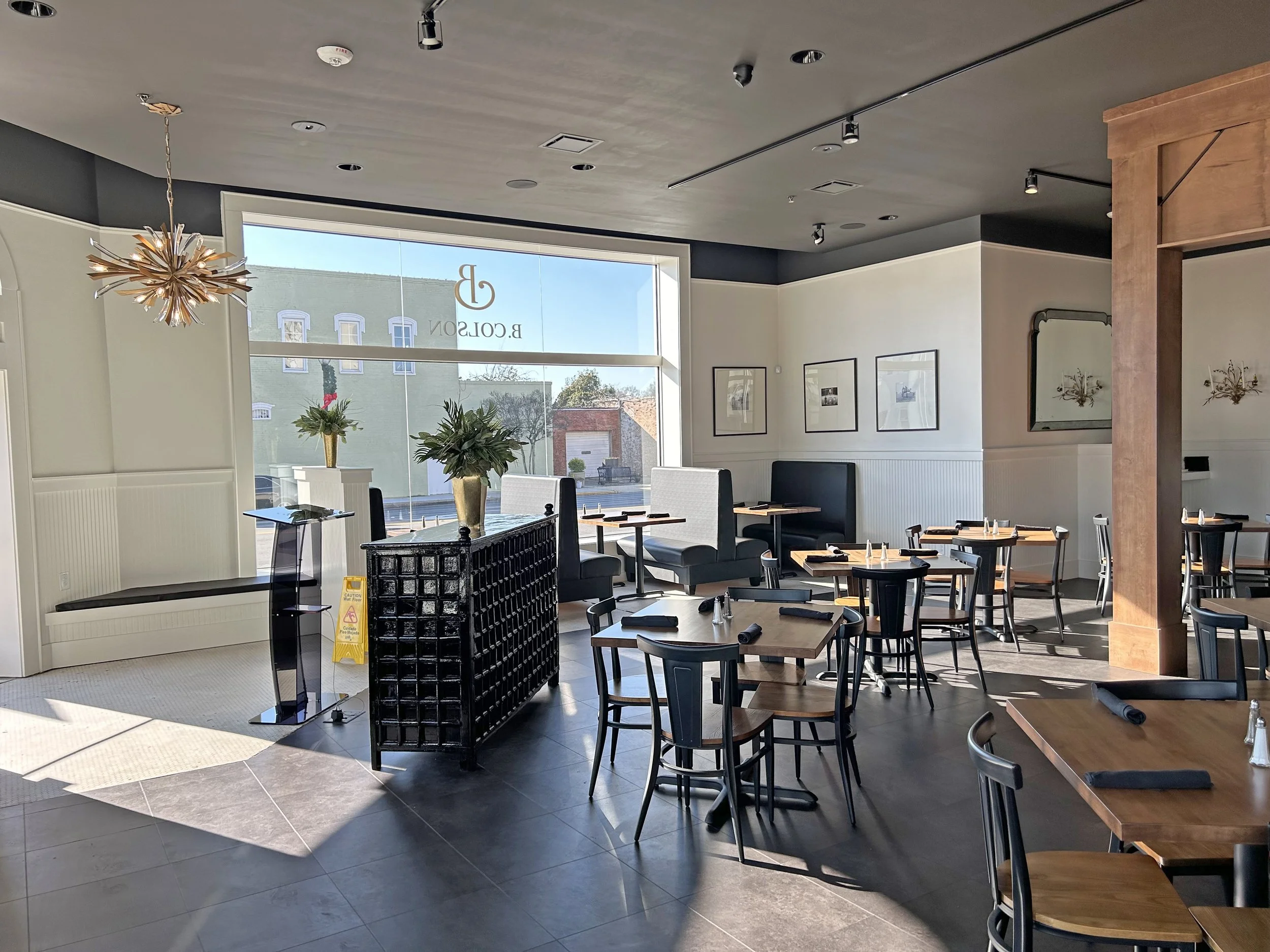 Interior of a modern restaurant with wooden and black furniture, large window, plants, framed artwork on white walls, and stylish lighting fixtures.