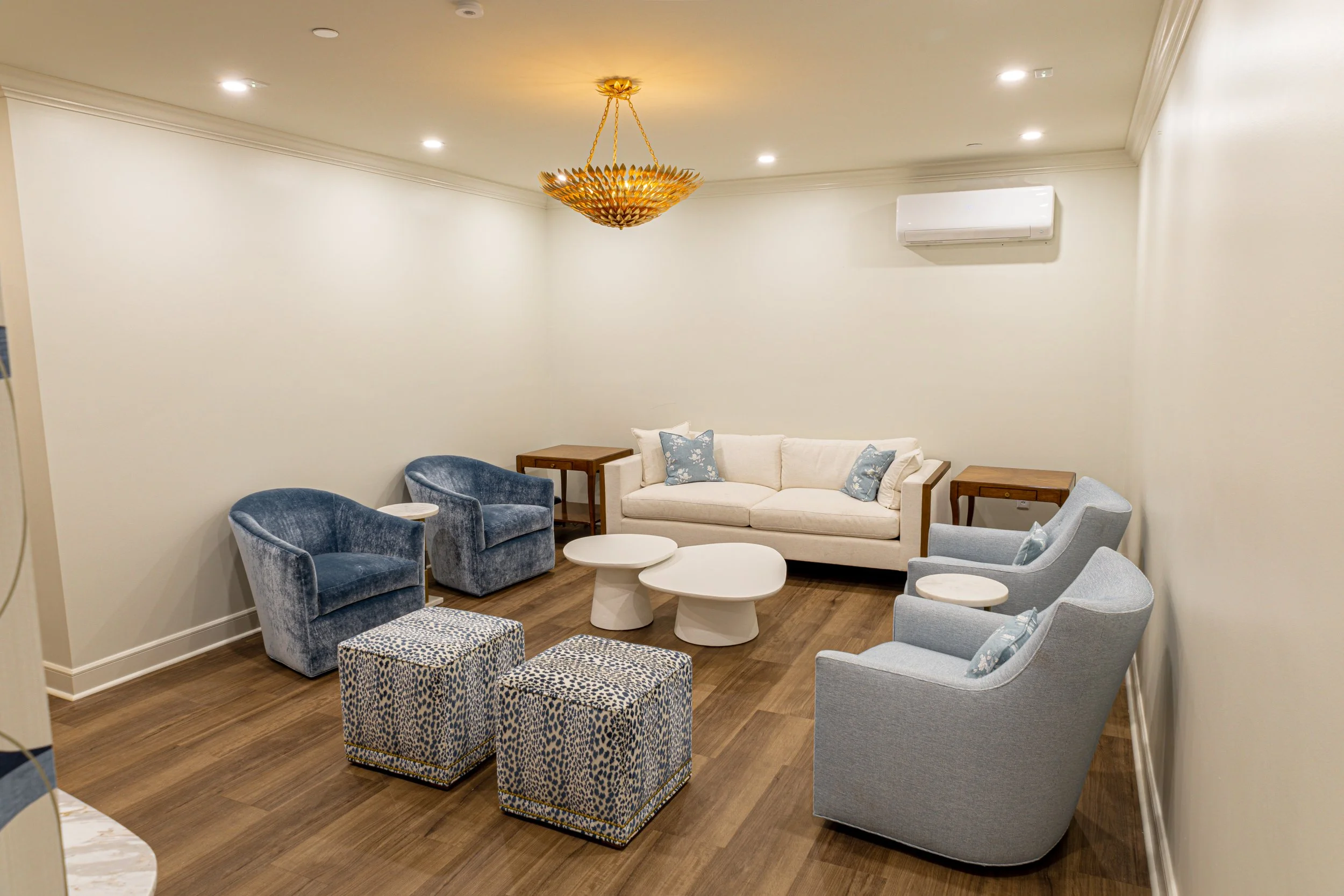 Living room with a white sofa with blue pillows, blue velvet armchairs, patterned ottomans, white coffee tables, wooden side tables, a decorative gold chandelier, and a white wall-mounted air conditioner.