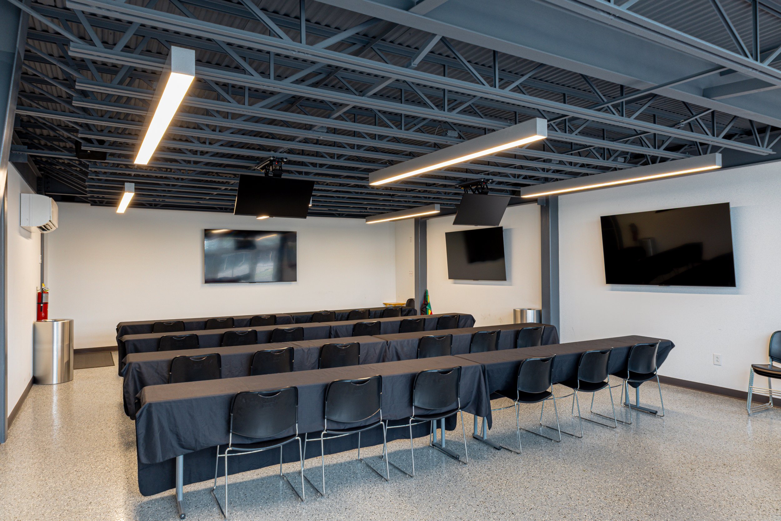 A conference room with rows of tables covered in black tablecloths and black chairs, large wall-mounted televisions, and modern lighting fixtures on a metal ceiling framework.