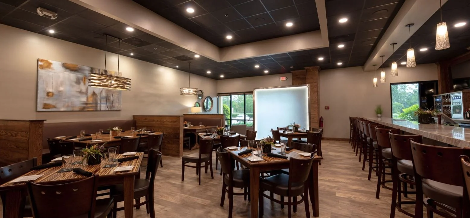 Interior view of a restaurant with wooden tables and chairs, modern light fixtures, and a bar area to the right, with windows showing greenery outside.