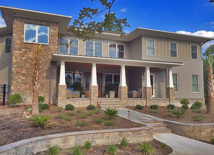 Front view of a modern two-story house with a stone and siding exterior, front porch with columns, and landscaped yard with small plants and trees.
