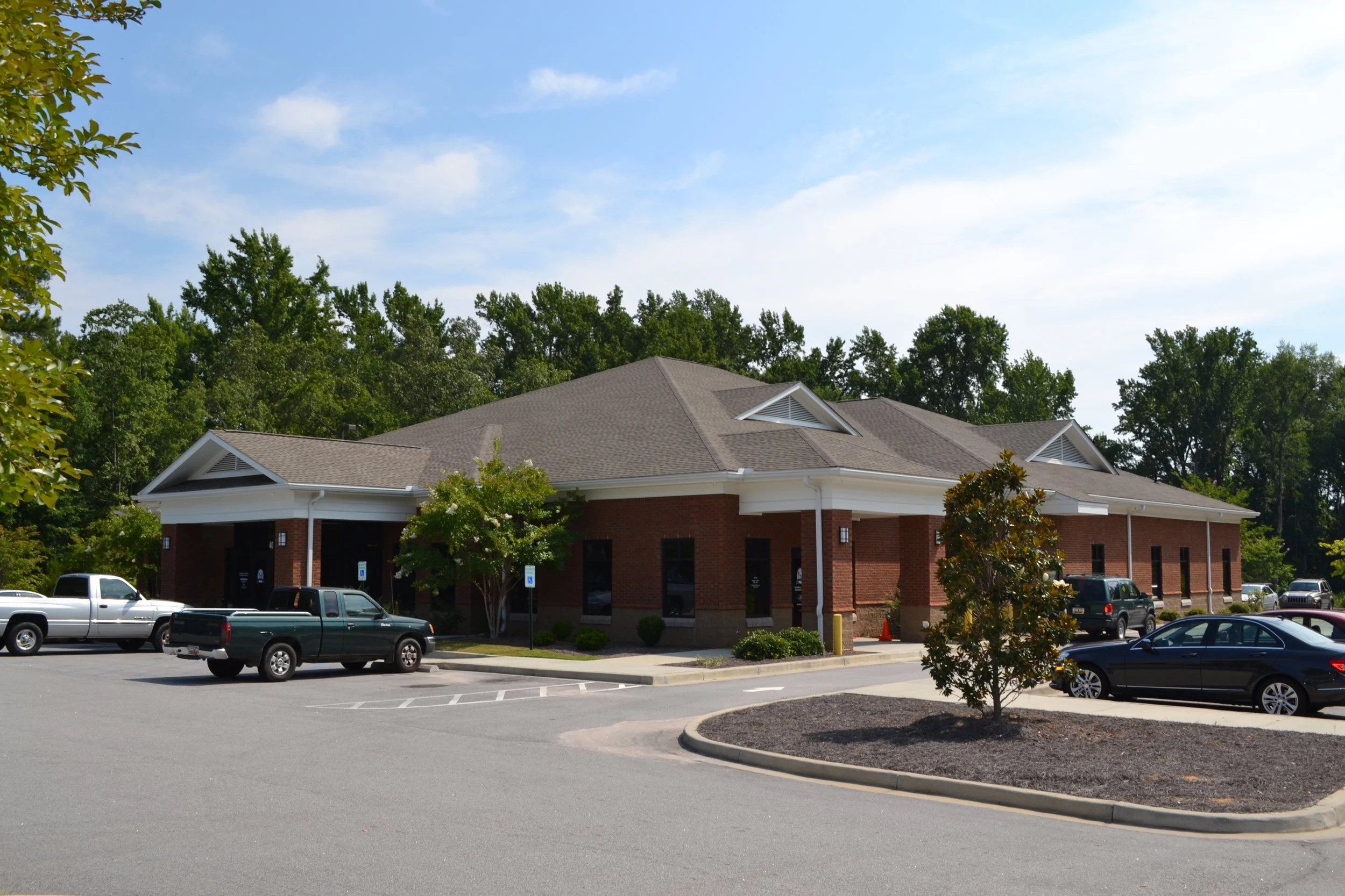 Parking lot in front of a brick building with a gray roof, surrounded by trees and parked cars under a blue sky with some clouds.