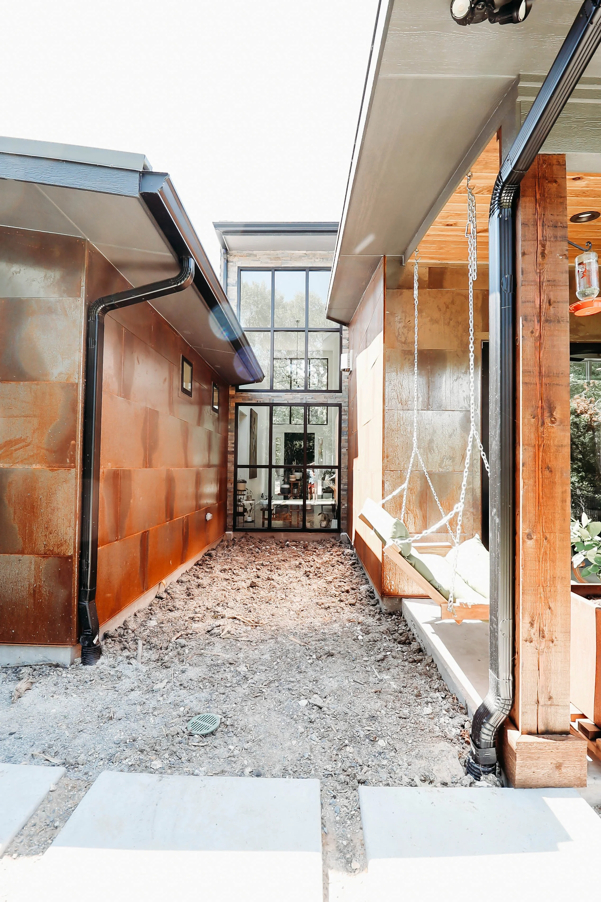 Construction site with a gravel ground, a foundation, and some stepping stones leading towards a glass door with a window above it. There are unfinished walls with wood and metal materials, and a swing with white cushions hanging from a hook. Black drainage pipes are installed along the walls.