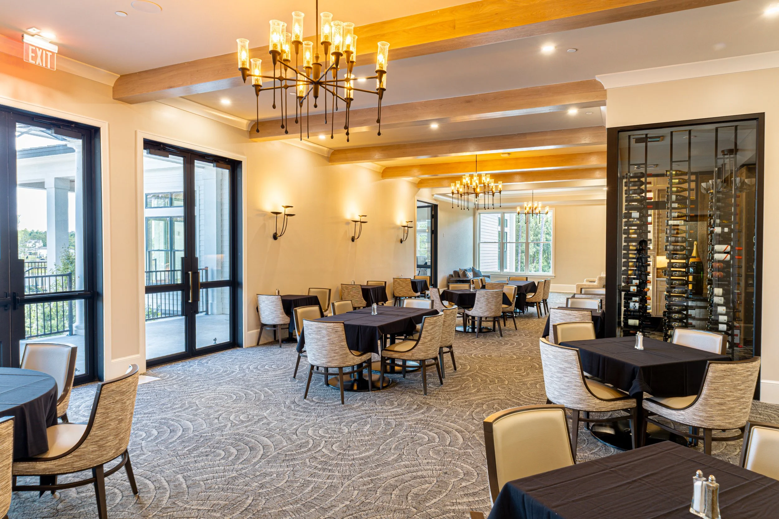 A spacious restaurant or event space with black tablecloth-covered tables and beige chairs arranged in rows. Large windows let in natural light, and there is a wine display on the right and ornate chandeliers hanging from the ceiling.