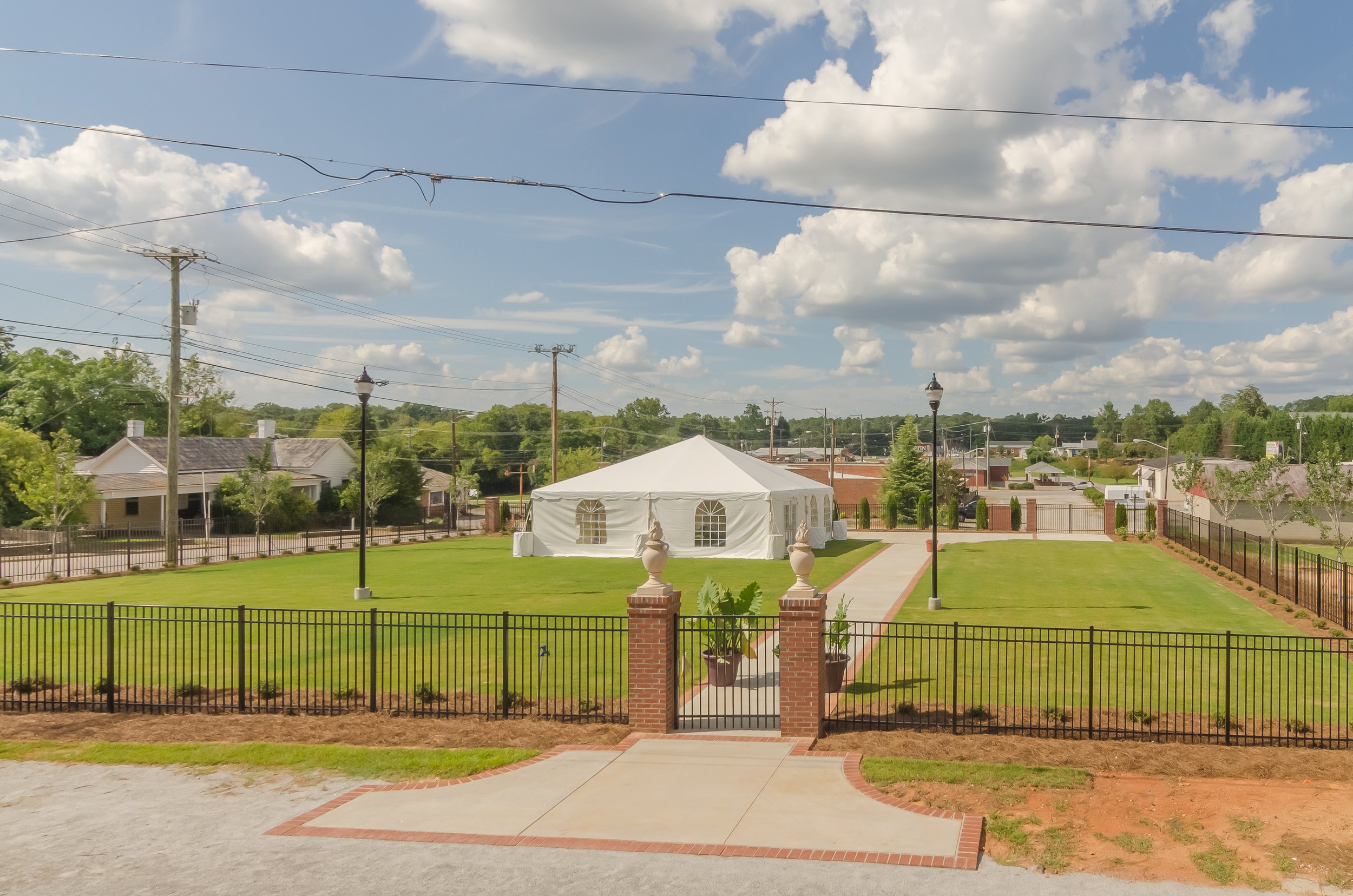 View of a backyard with a green lawn, a white tent, and a brick fence with decorative pillars. There are trees, houses, and power lines in the background under a partly cloudy sky.