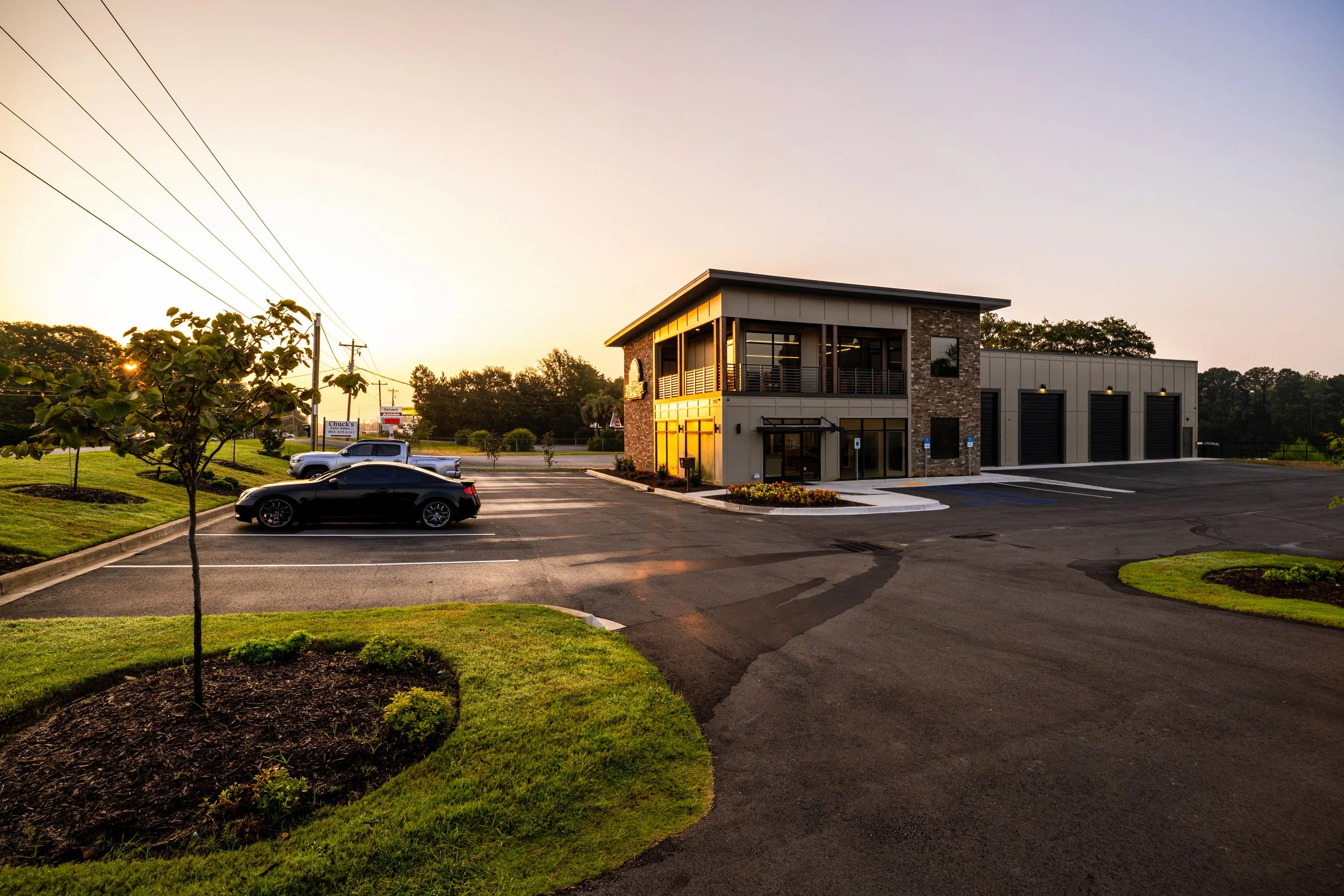 A modern two-story commercial building with a brick and metal exterior, large windows, and a small attached garage. The parking lot has a few cars, and the area is landscaped with grass, small trees, and bushes. The photo is taken at sunset with a cl