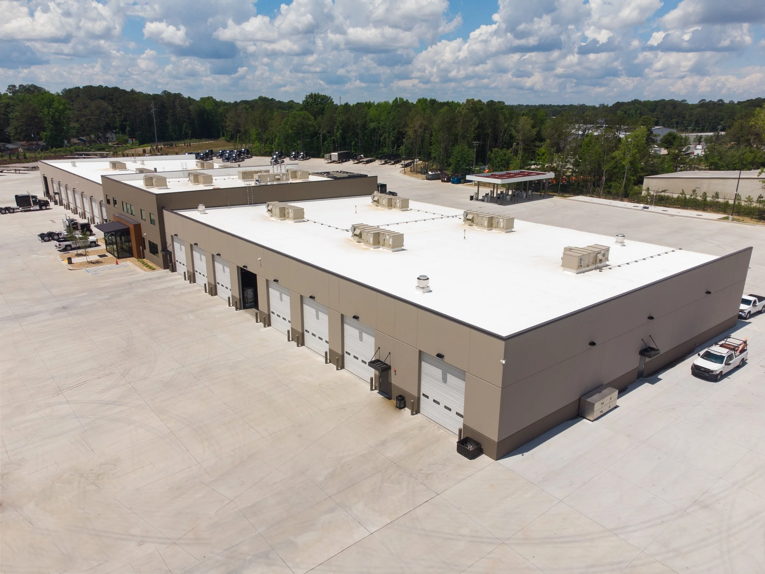 Aerial view of a large industrial building with a white roof, multiple garage doors, HVAC units, a parking lot with some trucks, and a vast empty paved area. Surrounding the building are trees and a partly cloudy sky.