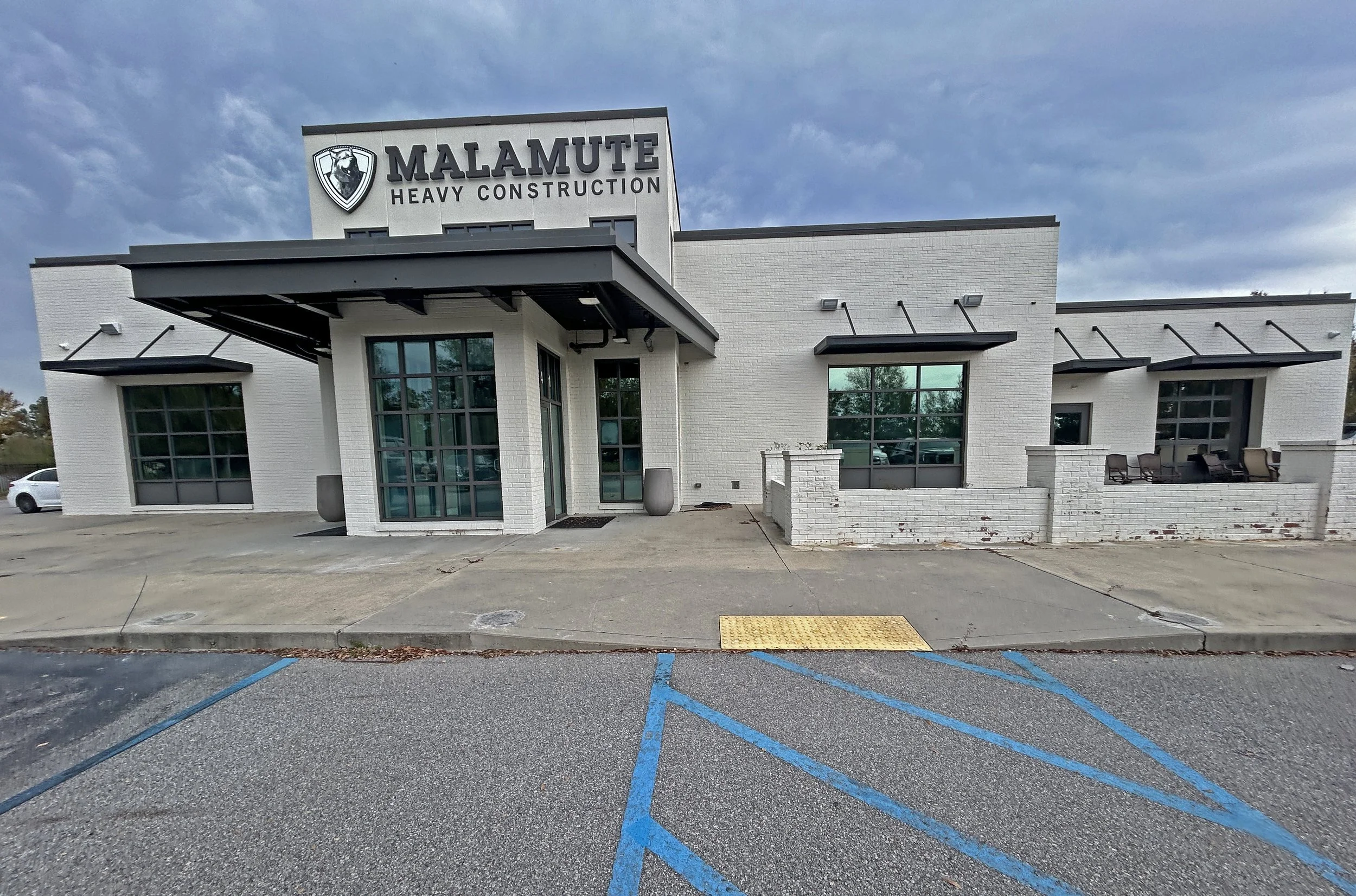 Exterior view of a white commercial building with signage for Malamute Heavy Construction, featuring a logo of a black and white dog. The building has large windows, a covered entrance, and is located in a parking lot with blue painted parking lines and a yellow textured ramp for accessibility. The sky is partly cloudy.