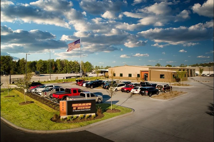 A parking lot outside a GMC dealership with a sign, several cars, a building, and an American flag flying in the wind under a partly cloudy sky.