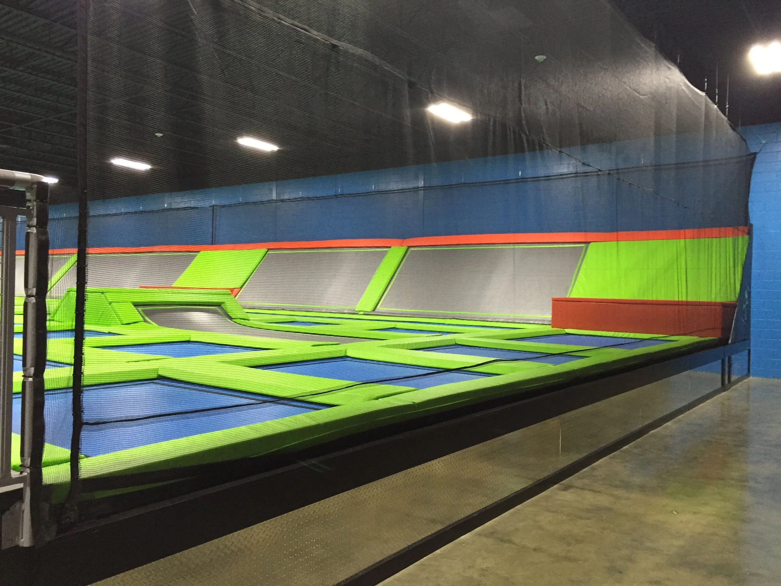 Indoor trampoline park with bright green, gray, blue, and red padded trampolines enclosed by black safety nets, illuminated by bright ceiling lights.