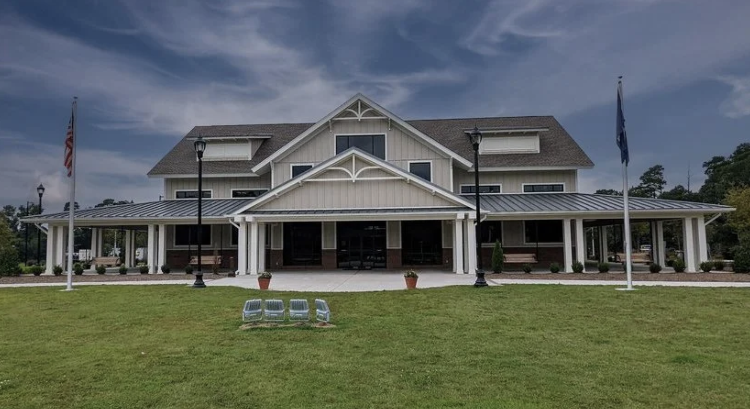 A large, multi-story building with a white exterior, gabled roof, and a covered porch area. There are flags on poles in front, and a green lawn with a paved walking path and benches. The sky above is partly cloudy.