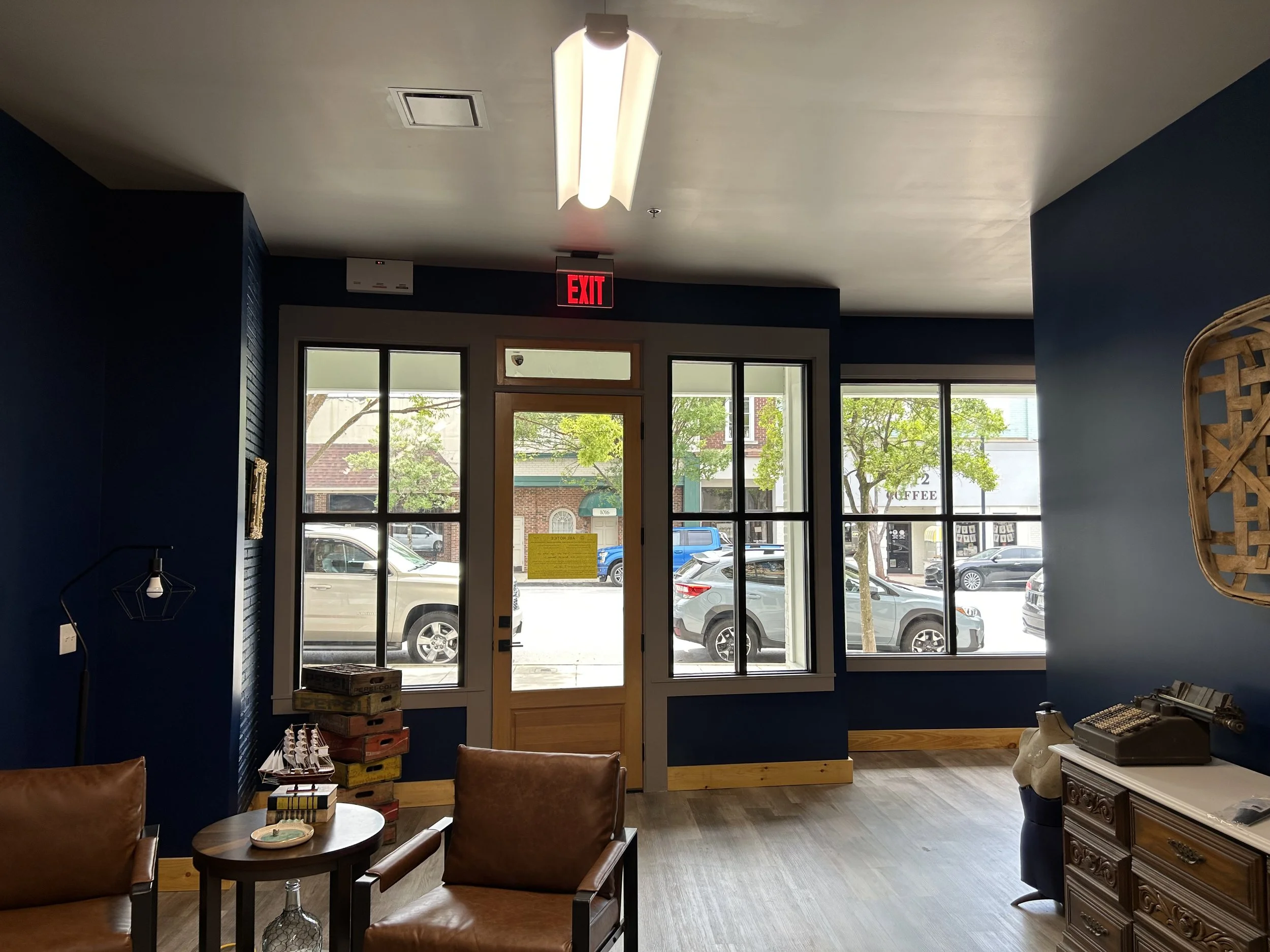 Interior of a coffee shop or cafe with large glass windows showing cars parked outside. Inside, there are leather chairs, a small round table, decorative items, and a wooden wall hanging. Fluorescent ceiling lights are visible, and a red 'EXIT' sign 