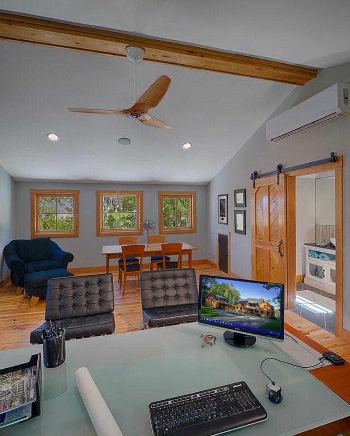 Home office with a desk, computer, and office chair in the foreground, a living area with a blue armchair, a dining table with four chairs, and three windows showing greenery in the background. There is a sliding barn door leading to a kitchen.